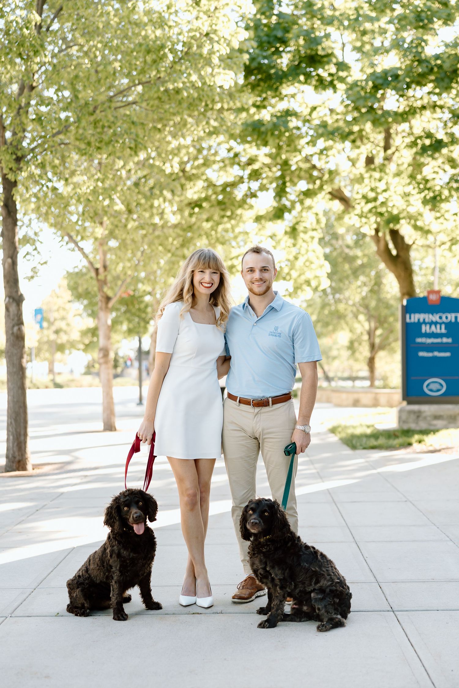 A couple stands with their black dog on a sunny sidewalk near Lippincott Hall in a series of casual outdoor portraits.
