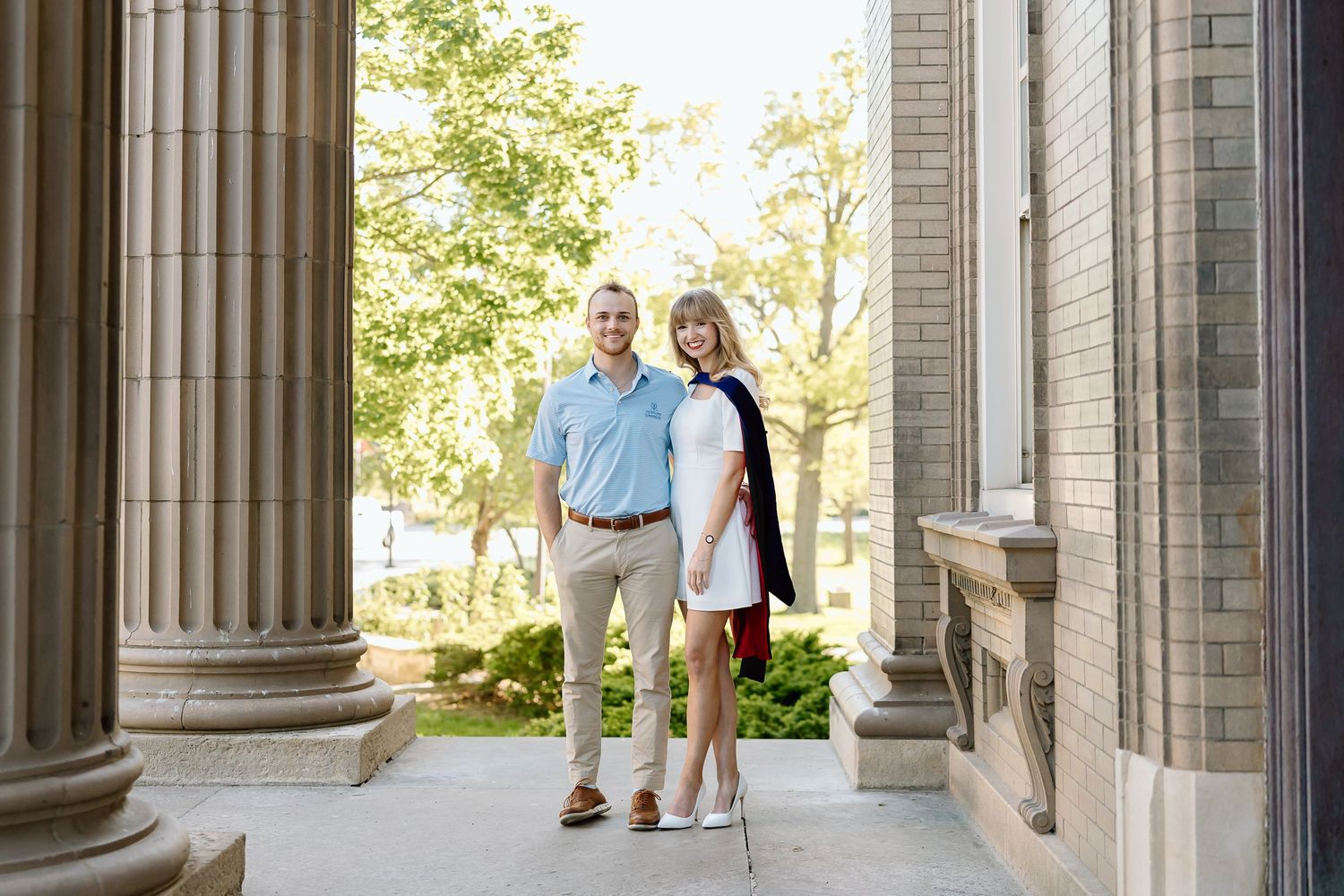 A couple stands between stone columns on a sunny day outside a classical building.