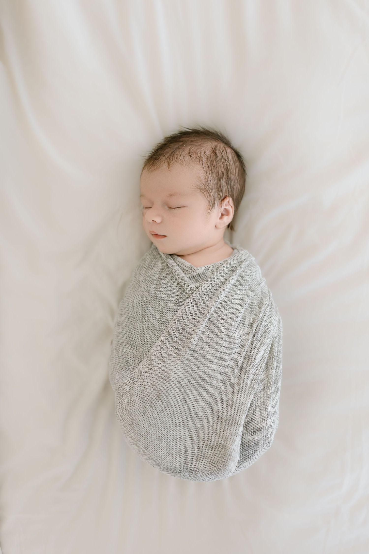 A sleeping newborn baby wrapped in a soft gray swaddle blanket lies on white bedding in a minimalist style photo series.