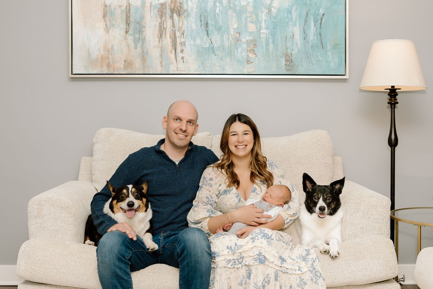 A couple sits on a white couch with their cats and newborn twins in a bright living room.