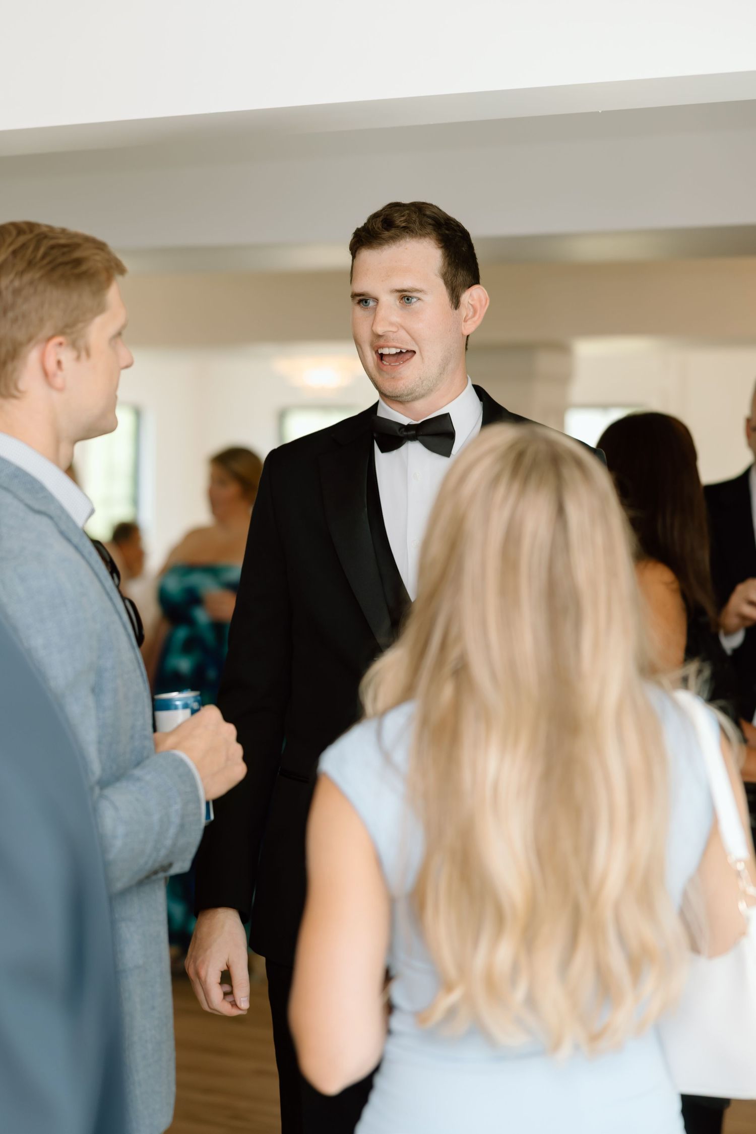 Groom in black tuxedo and bow tie conversing with wedding guests at a formal reception.