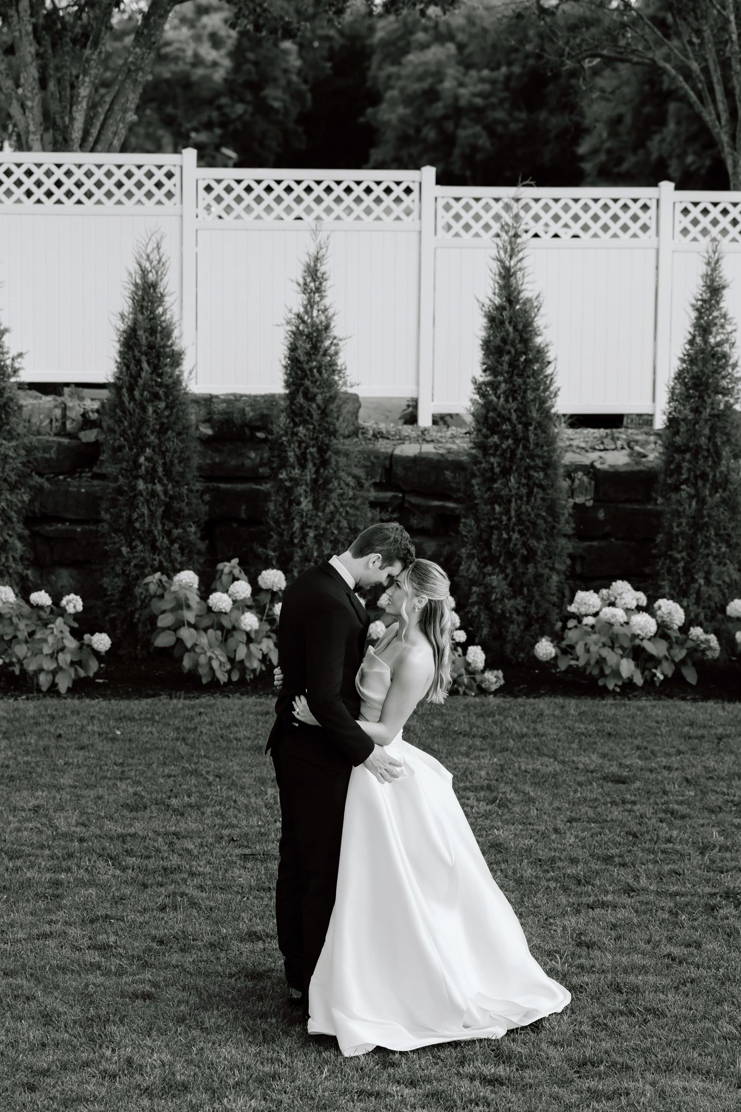Black and white photo of a bride and groom embracing on a lawn with tall evergreens and white lattice fence backdrop.