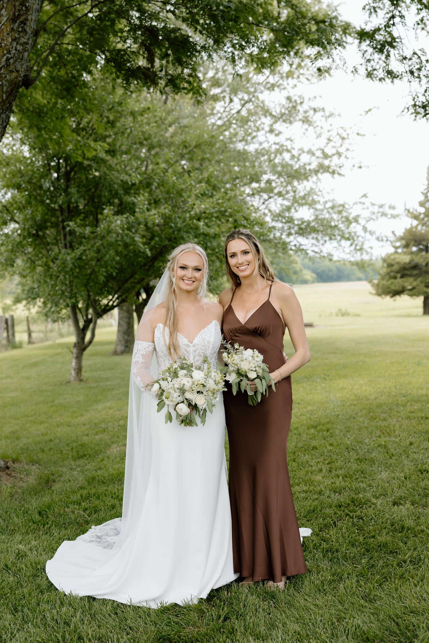 Bride in white gown and bridesmaid in brown satin dress holding bouquets outdoors on green lawn.