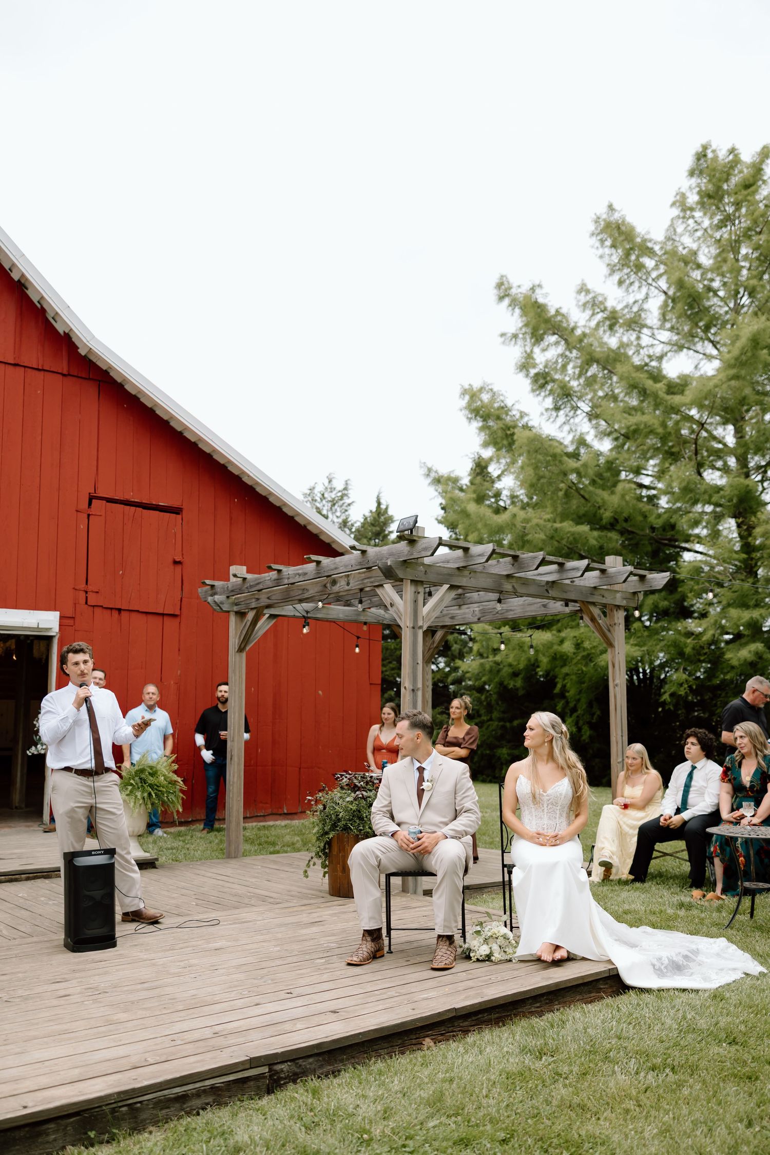 Outdoor barn wedding ceremony with couple seated on wooden deck, pergola, and red barn backdrop surrounded by green trees.