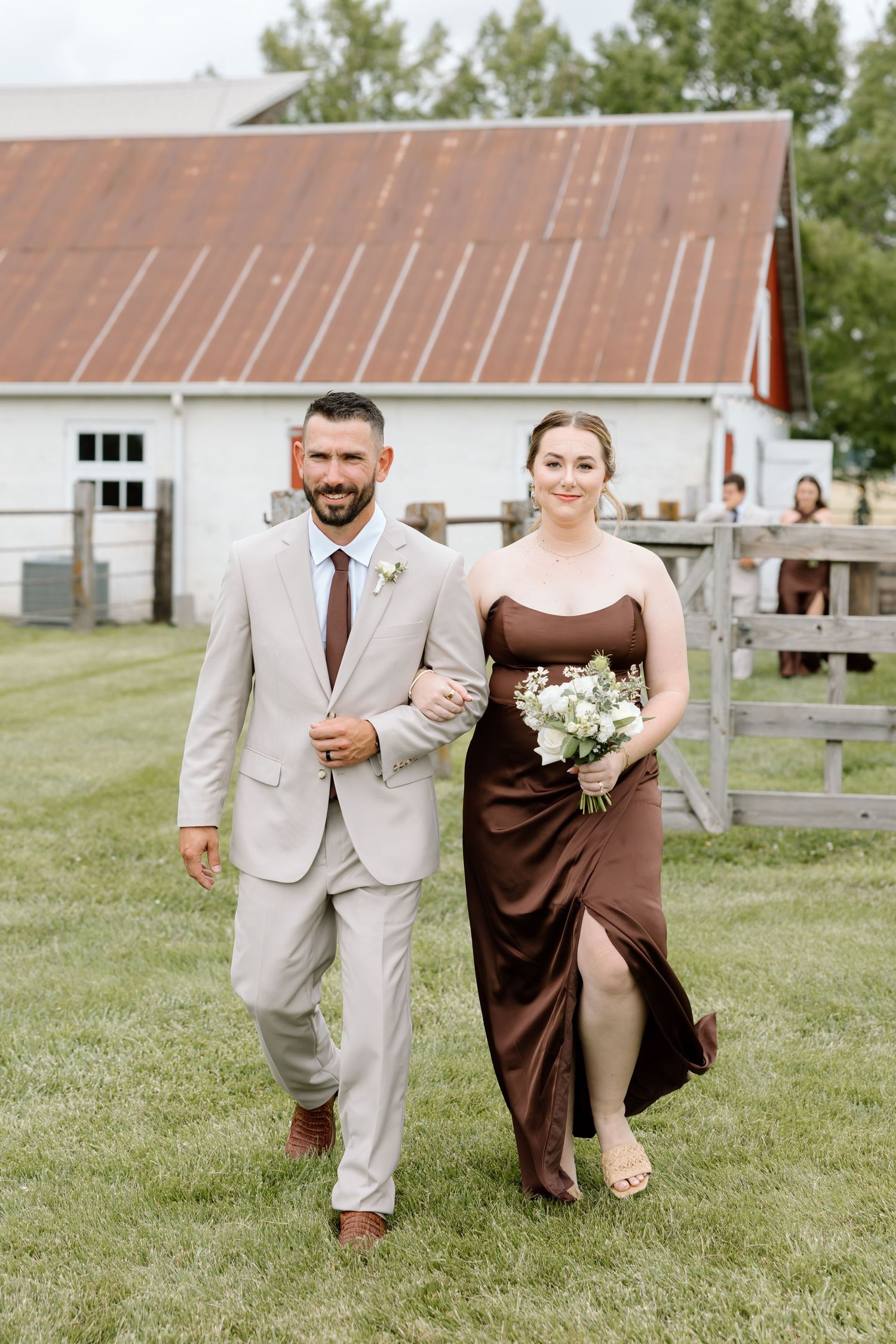 A groomsman in a tan suit escorts a bridesmaid in a brown satin gown across a green lawn at a barn wedding.