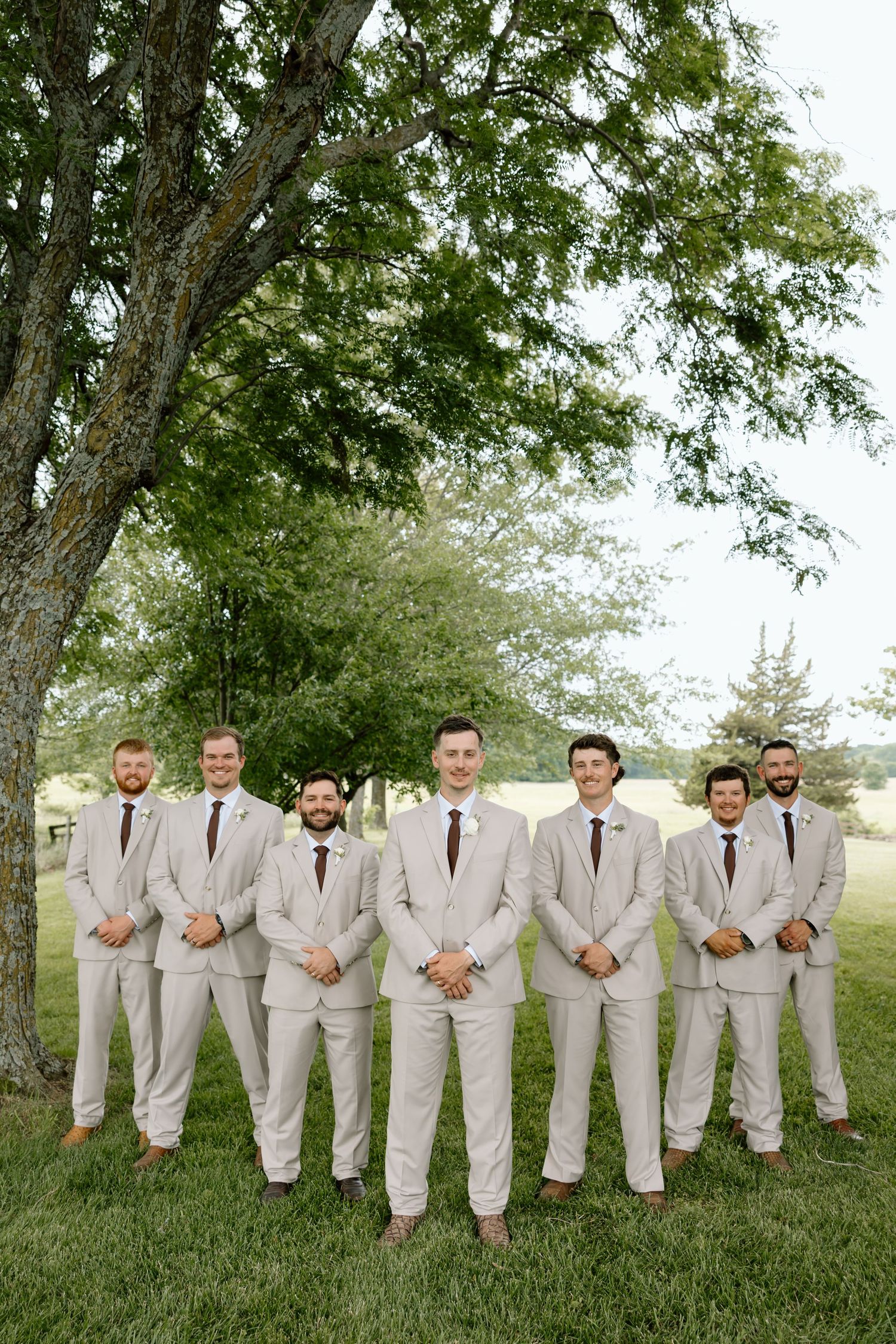 Groom and groomsmen in matching tan suits with dark ties posing outdoors under a large green tree on a wedding day.