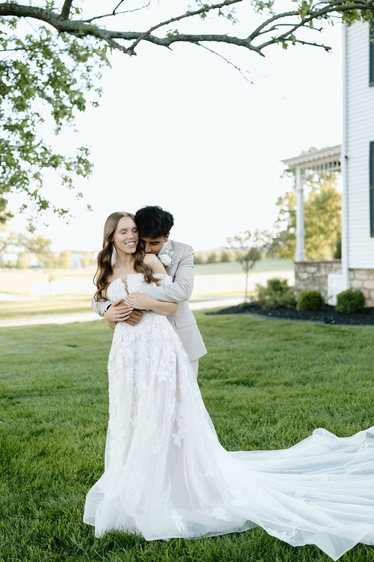 A smiling bride in a floral gown with a long train embraced by a groom in a tan suit outdoors.