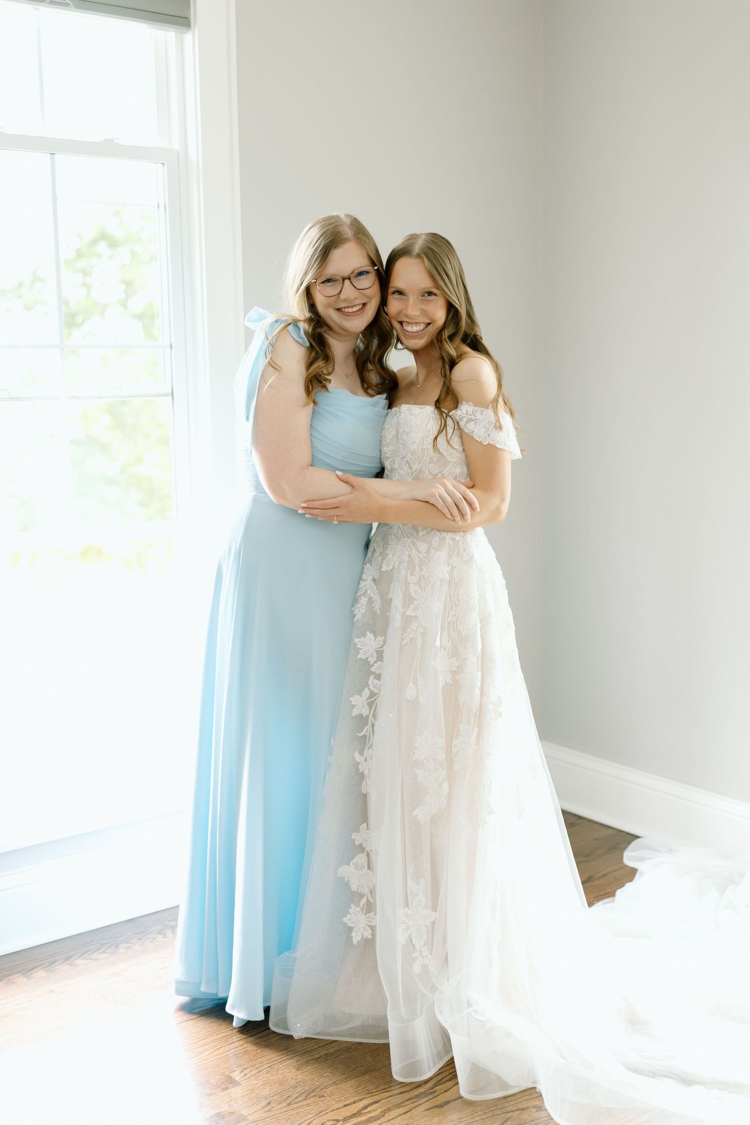 Bride in floral lace gown embraces bridesmaid in light blue dress, posing together near a bright window.