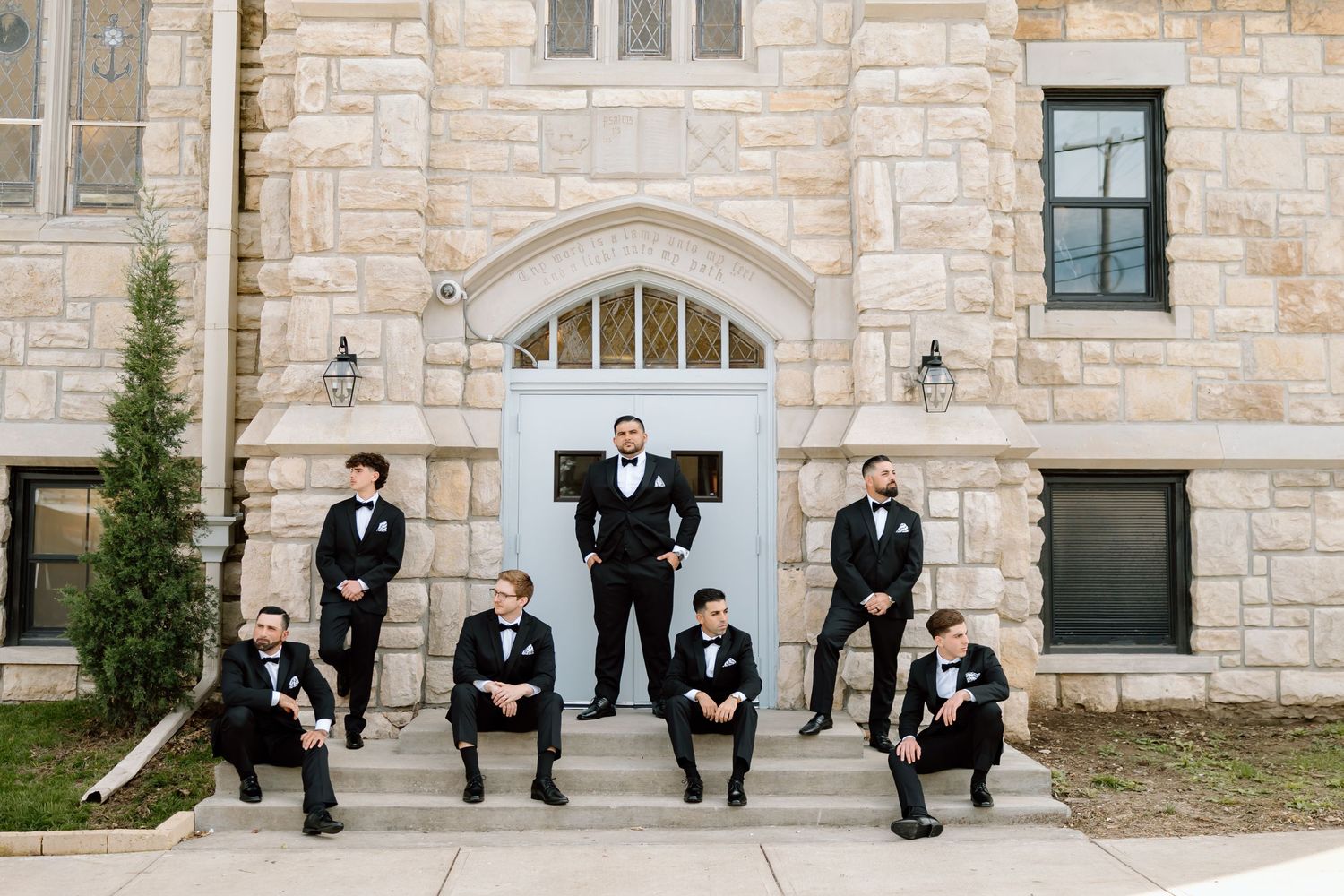 Groom stands centered on church steps flanked by six groomsmen in black tuxedos and bow ties posing for a wedding photo.