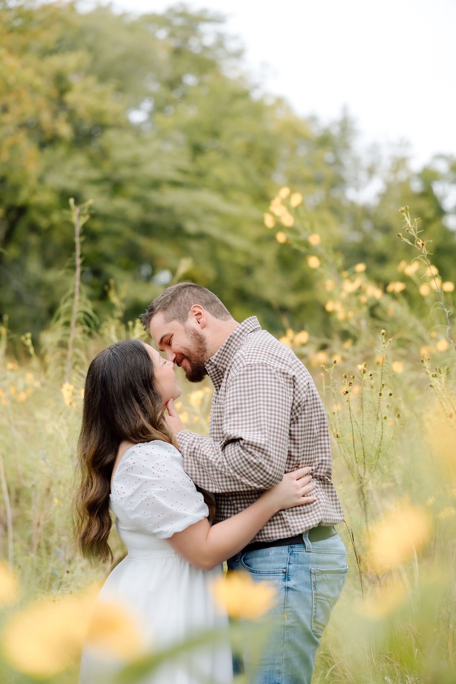 A couple shares a romantic embrace in a field with yellow flowers and trees in the background.