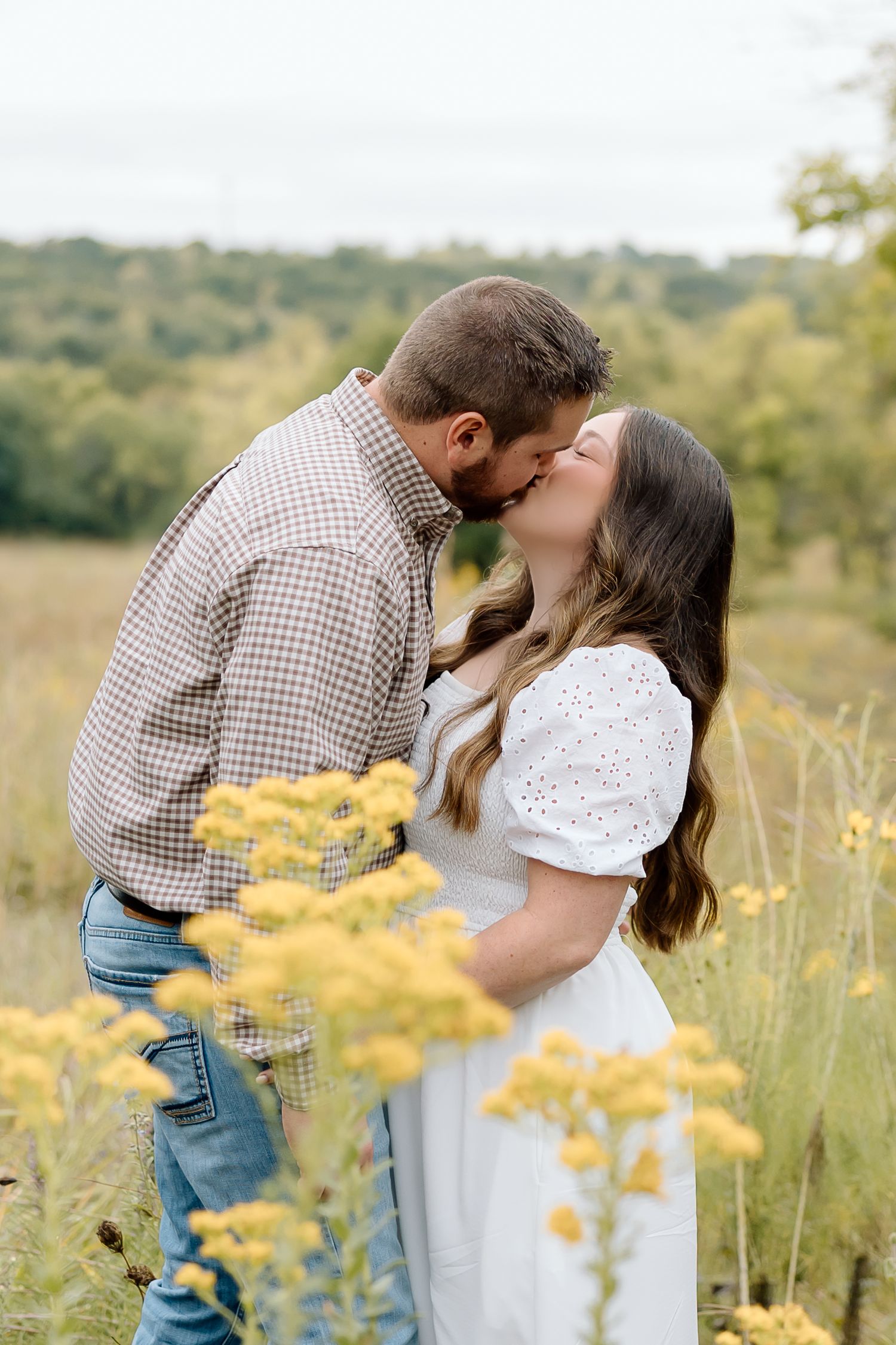 Intimate kiss shared between couple surrounded by yellow wildflowers in countryside setting.