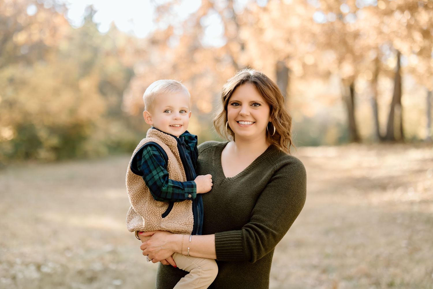 An autumn family portrait features a parent holding a small child against a backdrop of fall foliage.