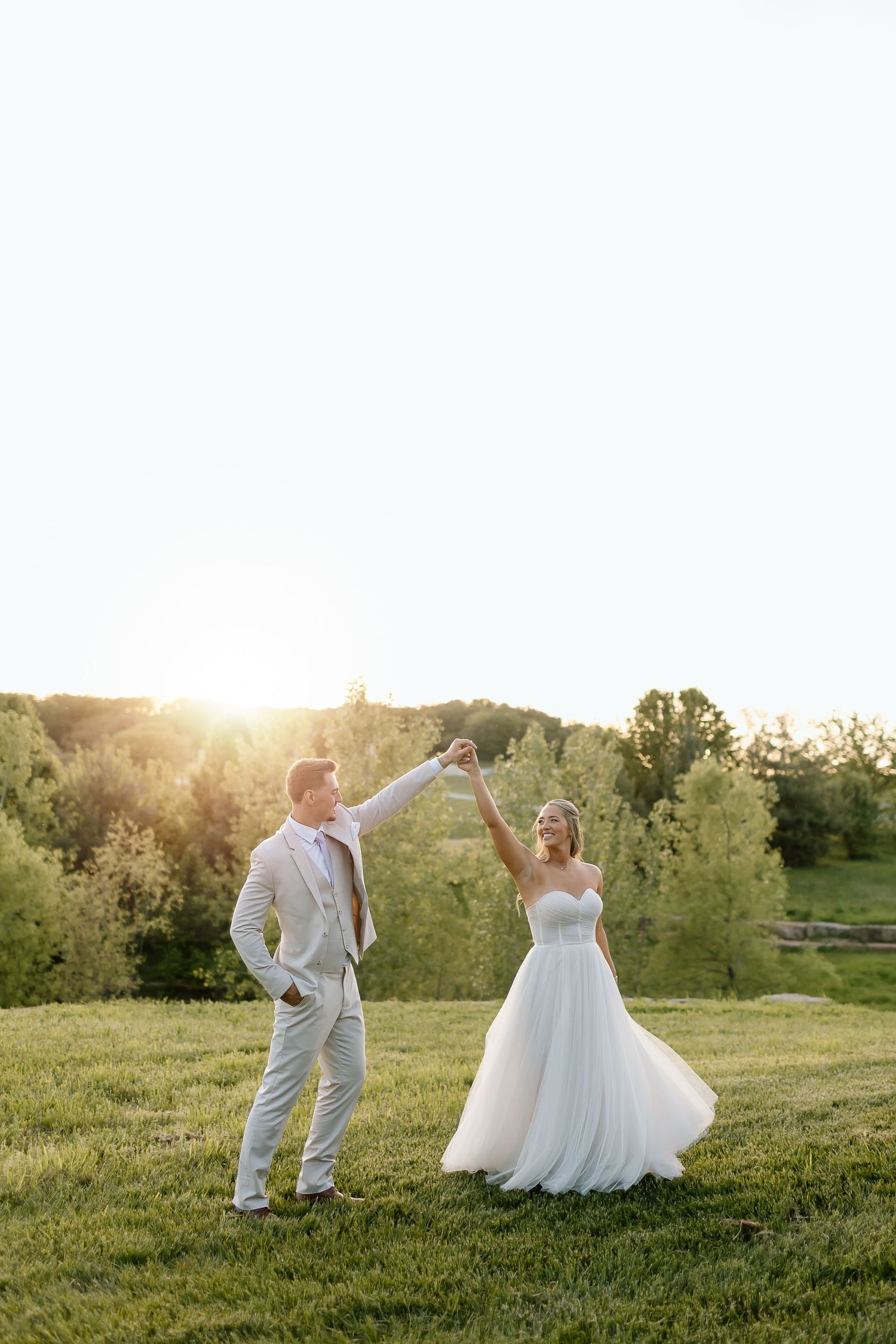 Bride twirls in her dress while groom watches during sunset photo session.