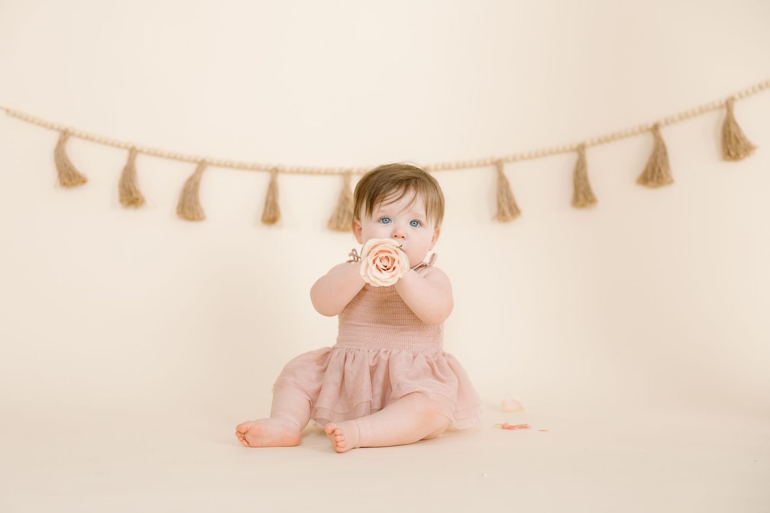 A small child in a pink dress sits under a tassel garland against a cream backdrop.