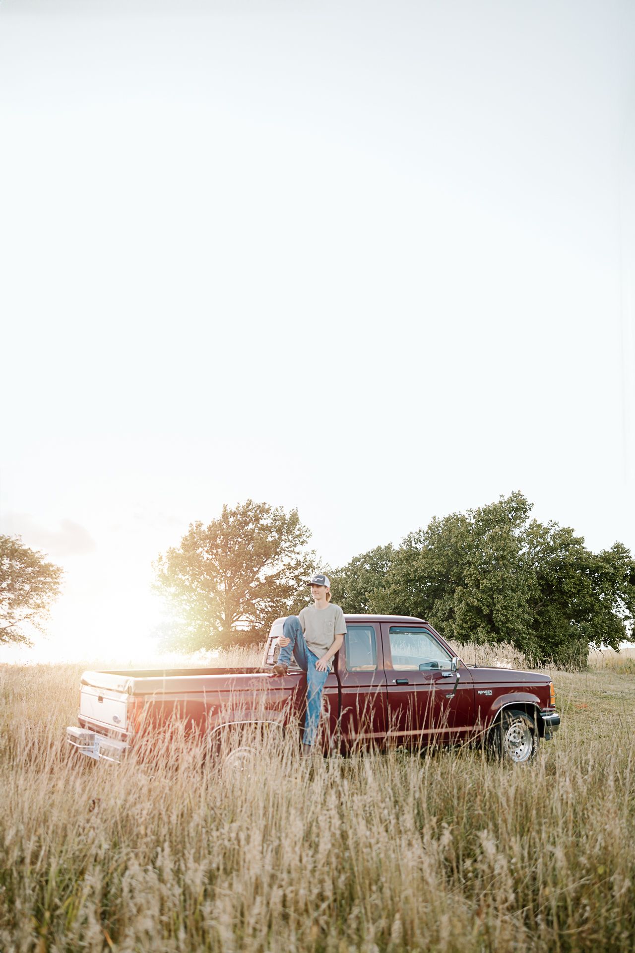 A vintage red pickup truck parked in a sunlit field of tall grass with trees in the background.