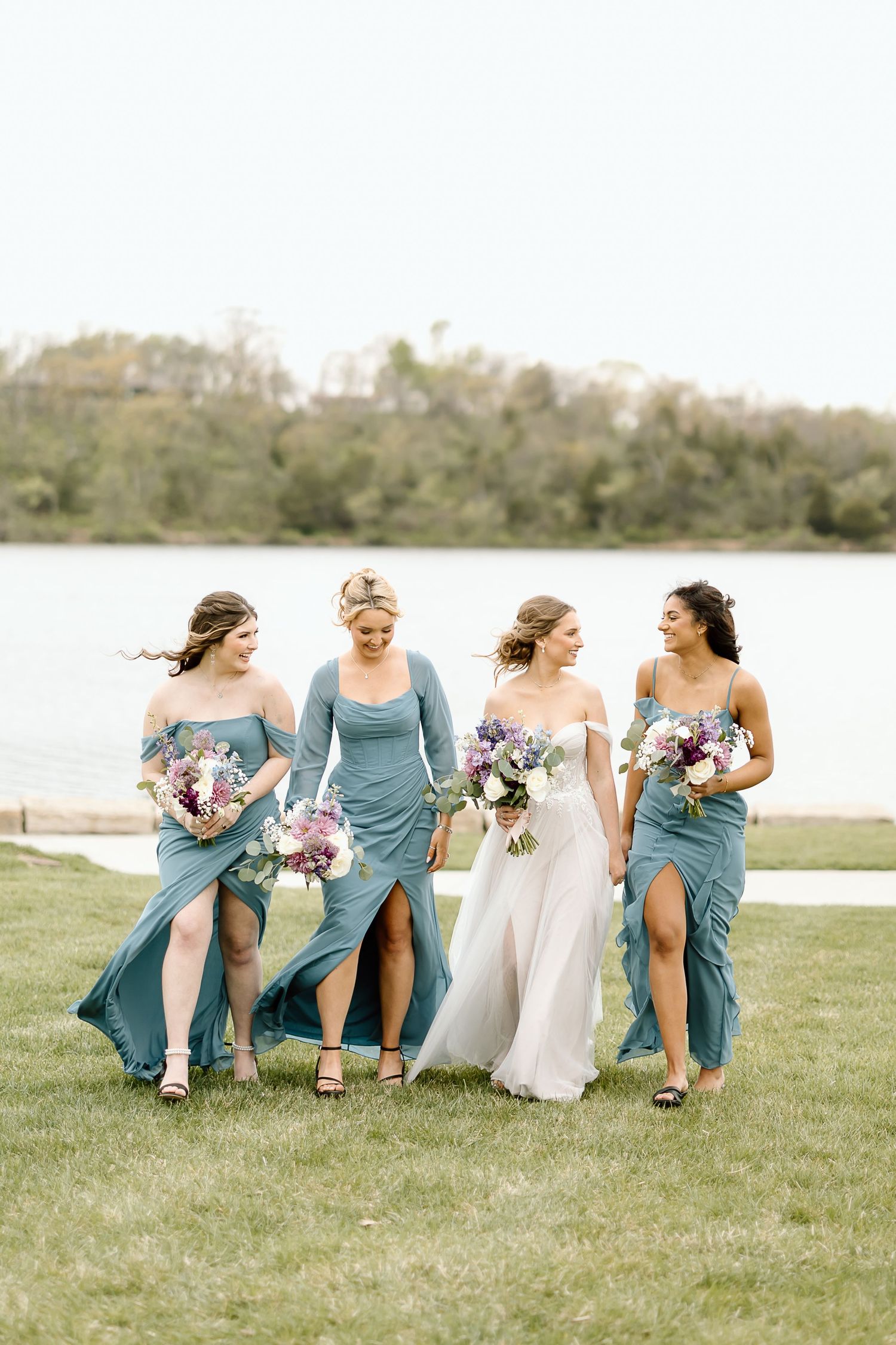 Bridesmaids in flowing dusty blue dresses walk together on grass by a lake.
