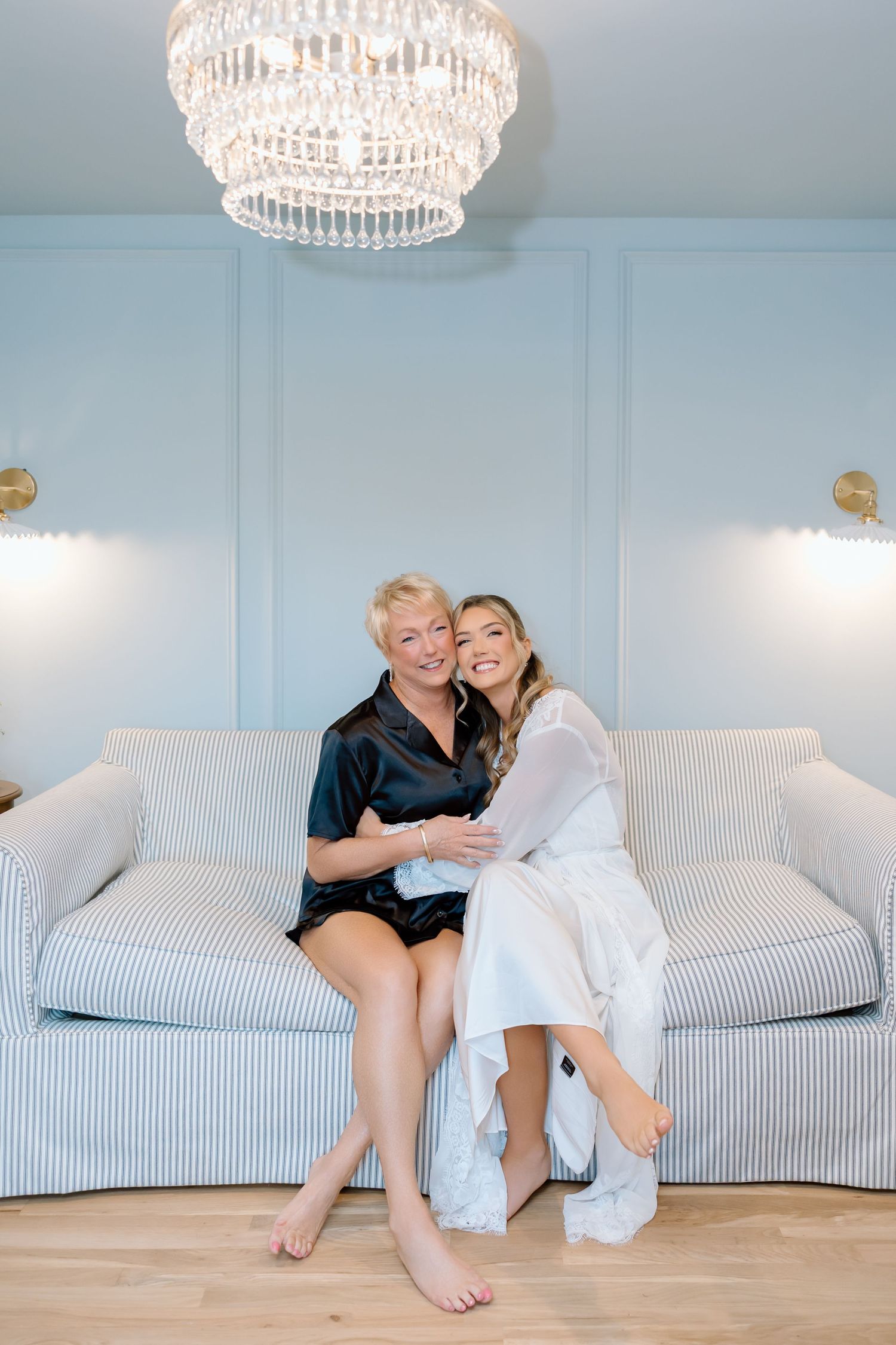 Two women in silk robes smile together on a striped sofa beneath a crystal chandelier in a blue room.