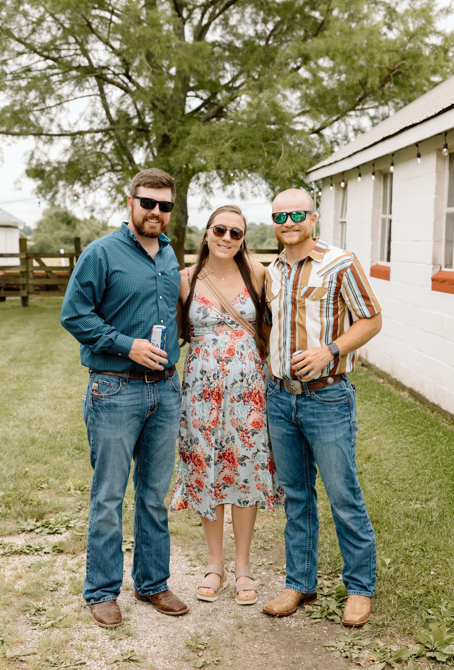 Three friends in sunglasses and jeans pose outdoors at a country gathering, woman in floral dress flanked by two men.