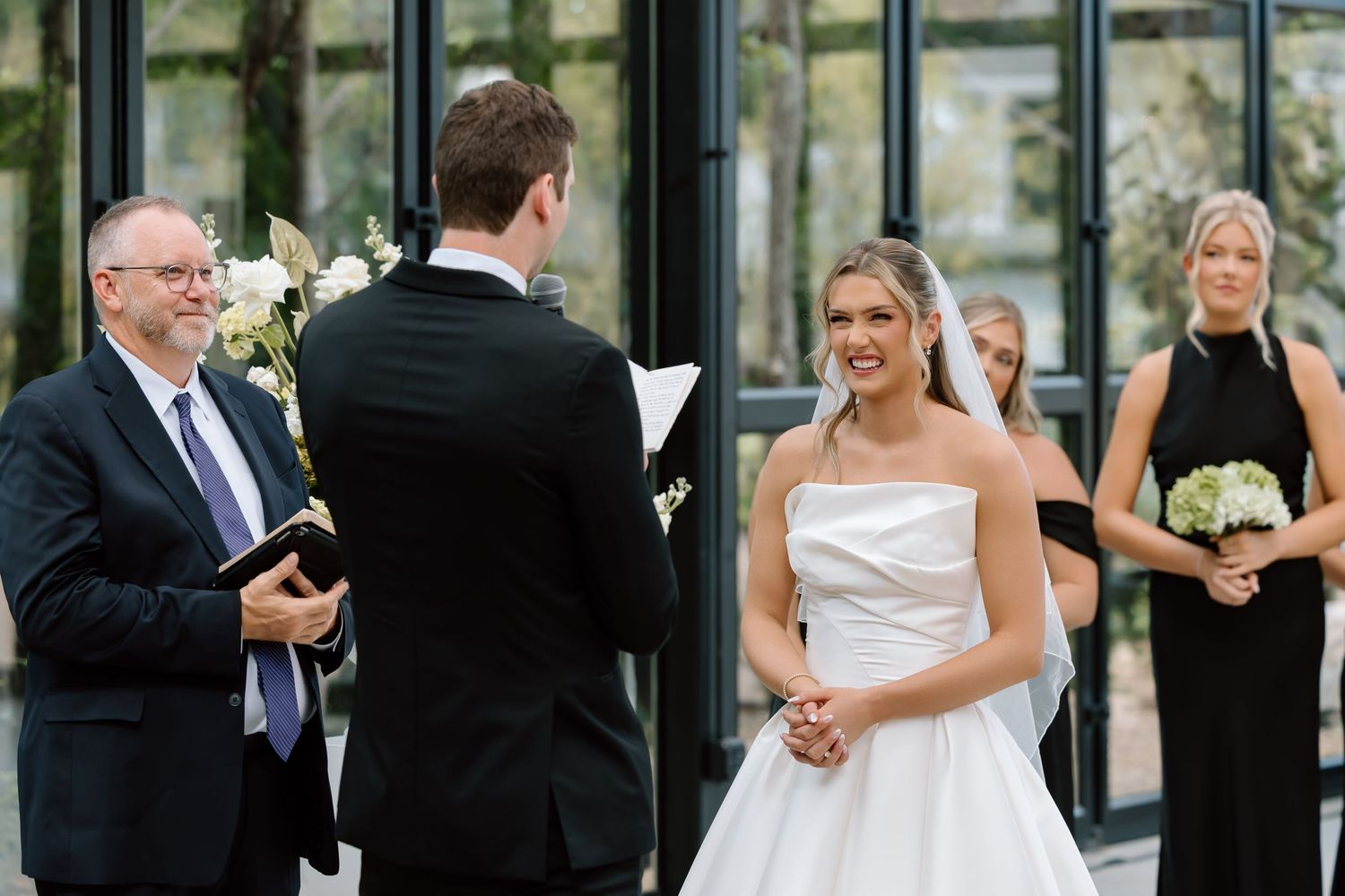 Bride smiling joyfully at groom during outdoor wedding ceremony with officiant and bridesmaid in elegant black dress.