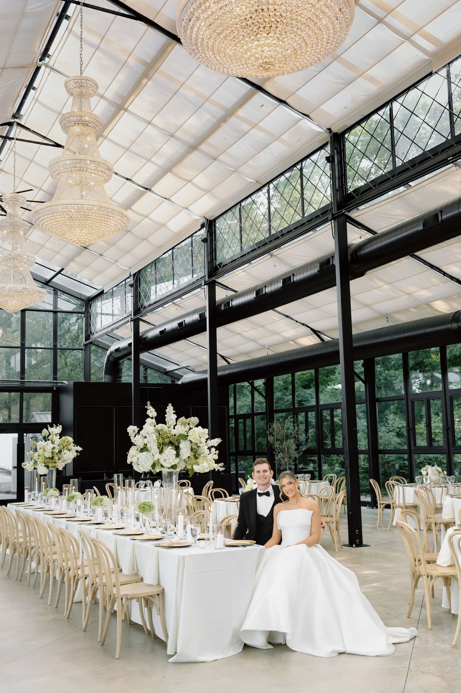 Bride and groom seated at elegant wedding reception table in glass greenhouse venue with crystal chandeliers.