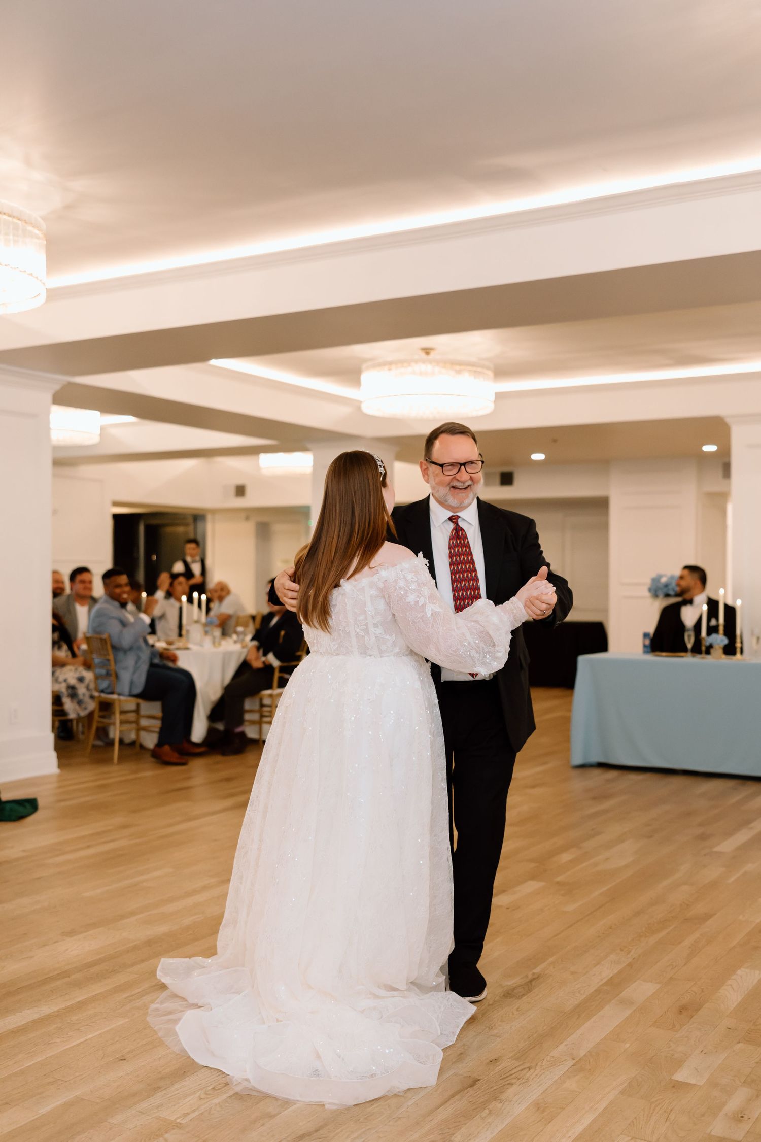 Bride and father share first dance at elegant wedding reception with guests seated at tables in background.