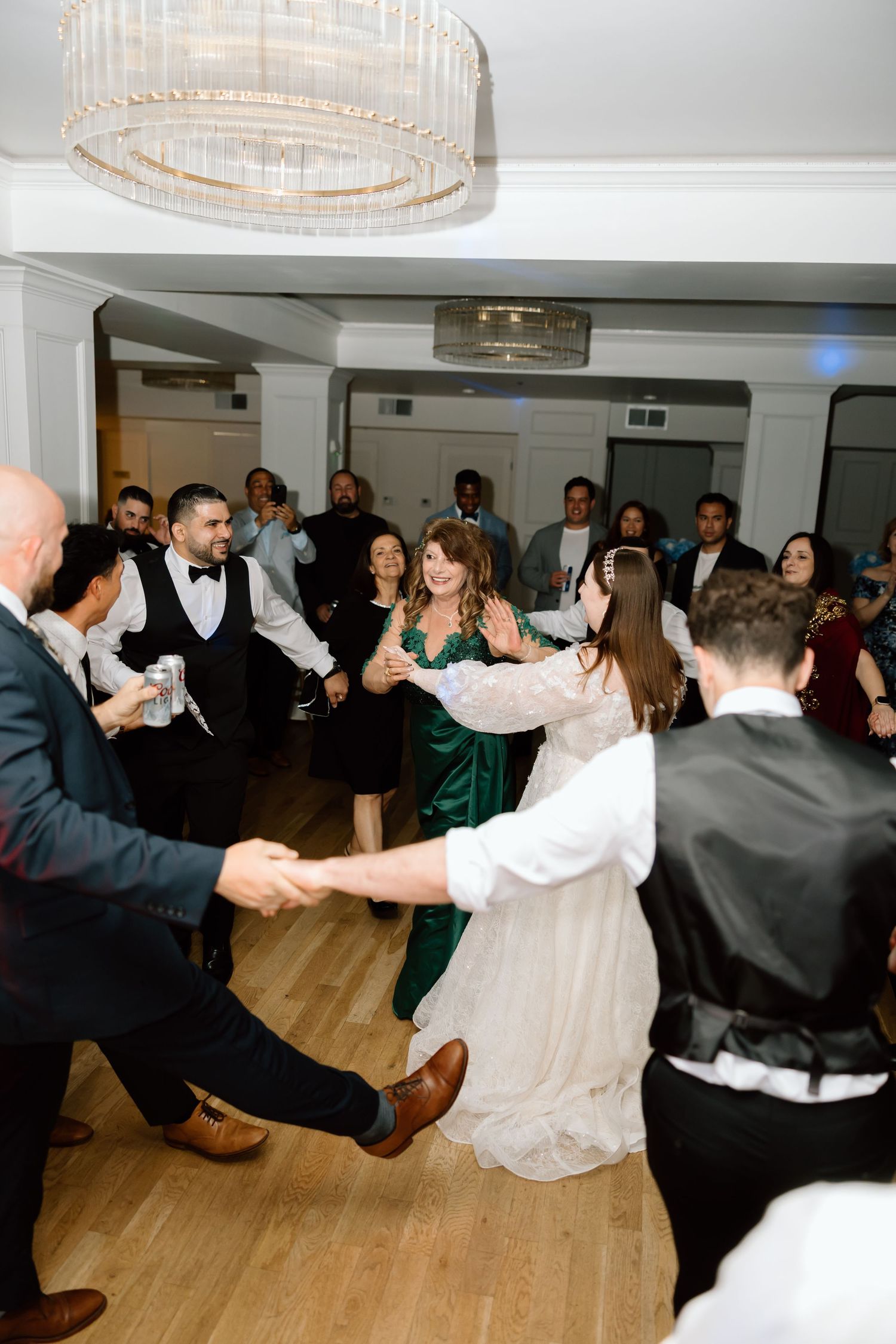 Wedding guests joyfully dancing in a circle at a reception venue with elegant chandeliers overhead.