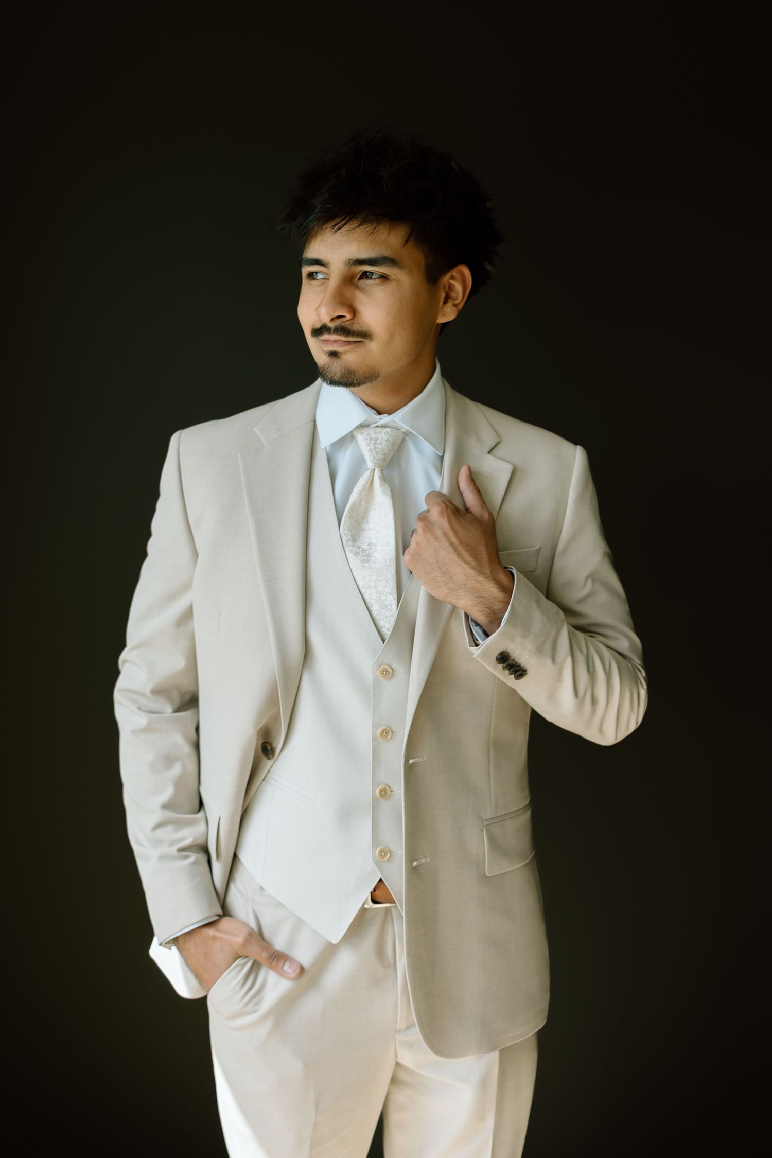 Elegant man in cream suit with light blue tie and vest poses confidently against dark background.