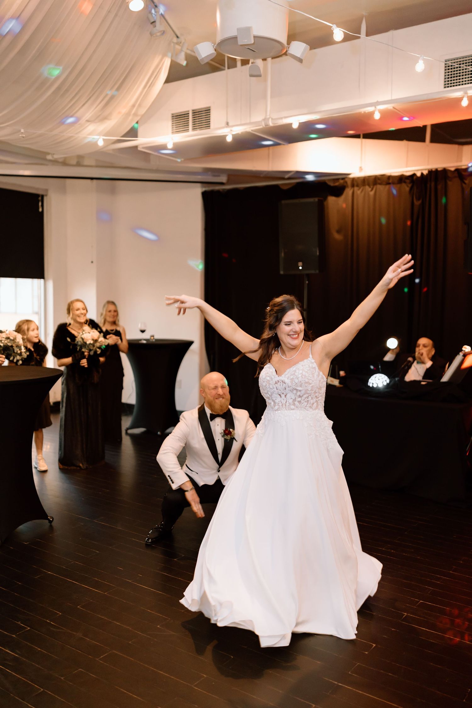 A couple dances together at a wedding reception on a dark hardwood floor under draped fabric and spotlights.