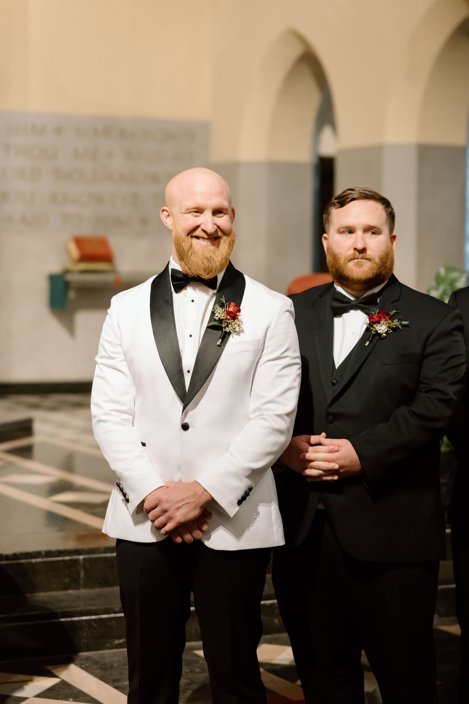 Two people in formal wedding attire, one wearing a white dinner jacket and one in black, smile together at a church.