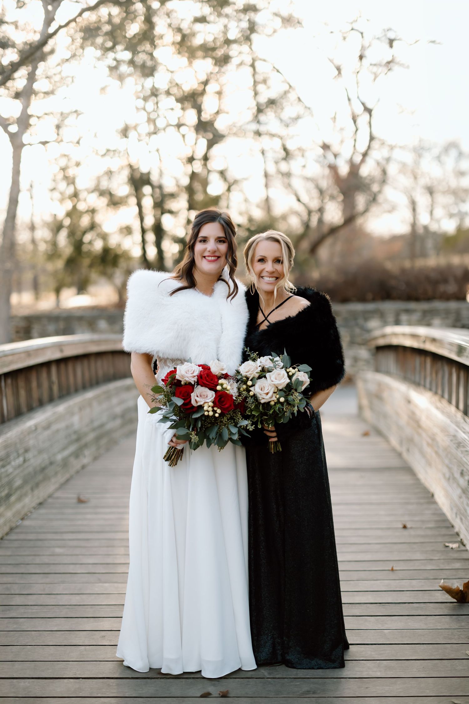Two people in contrasting white and black dresses stand together on a wooden bridge holding floral bouquets.