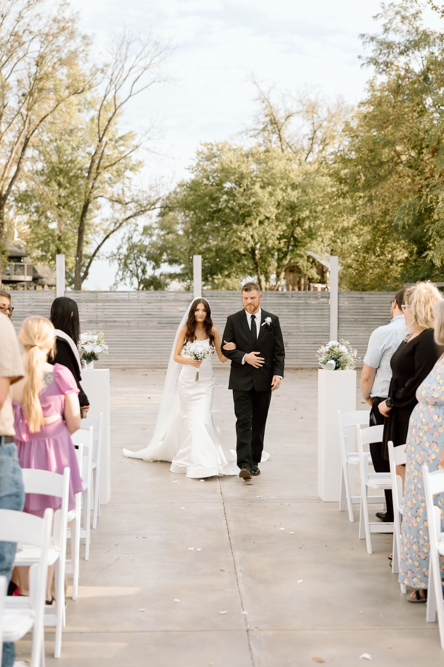 Wedding guests seated in white chairs outdoors during a socially distanced ceremony with trees in the background.