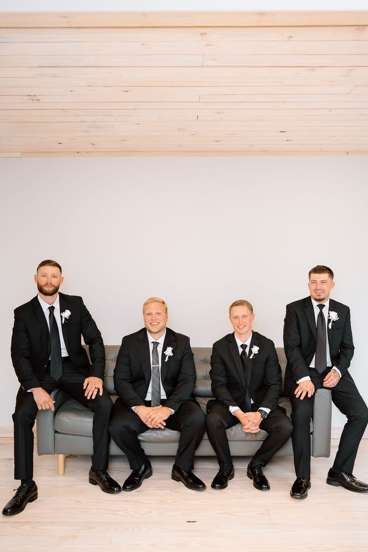 Four groomsmen in black suits and ties sit together on a gray couch against a white wall with wood trim.