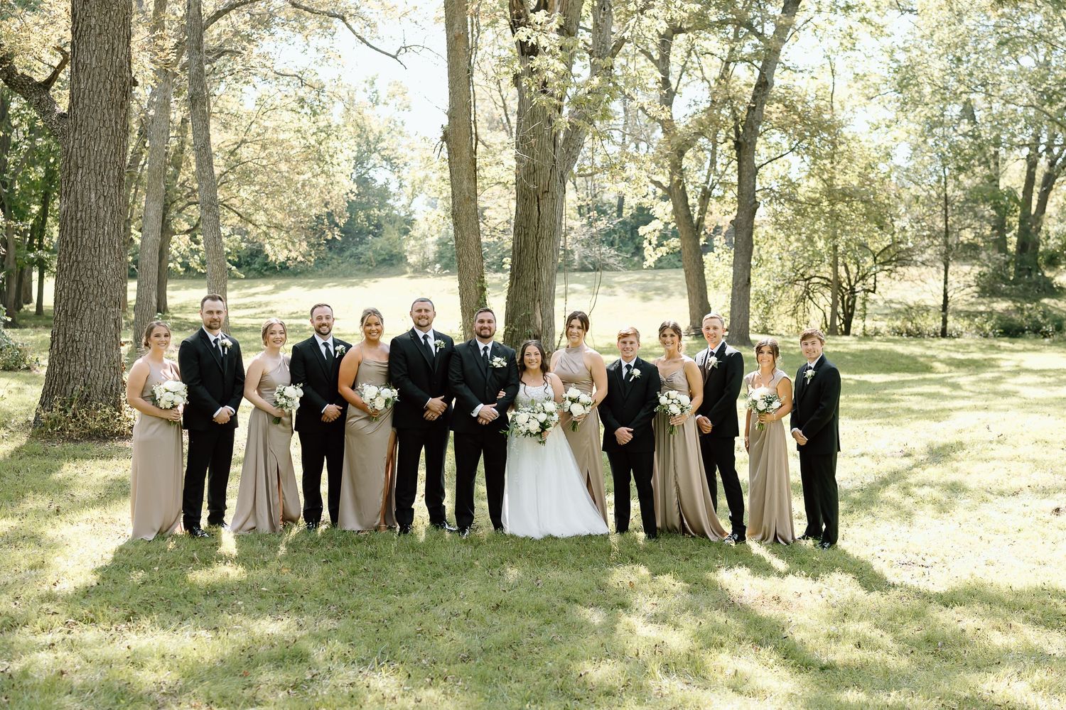 Wedding party members stand in a row under tall autumn trees during an outdoor ceremony.