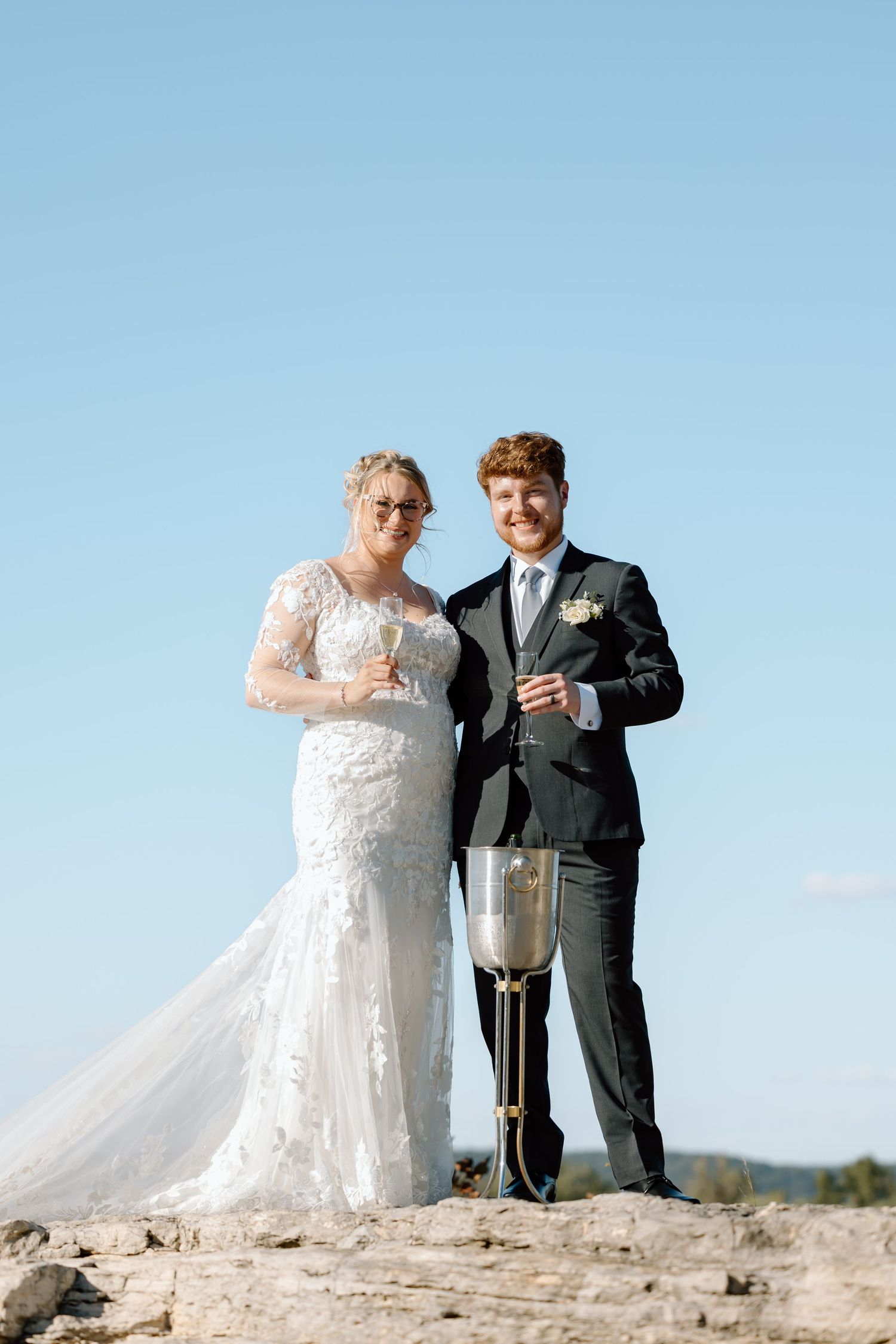 A couple in wedding attire pops champagne together while standing on a rocky outcrop against a blue sky.