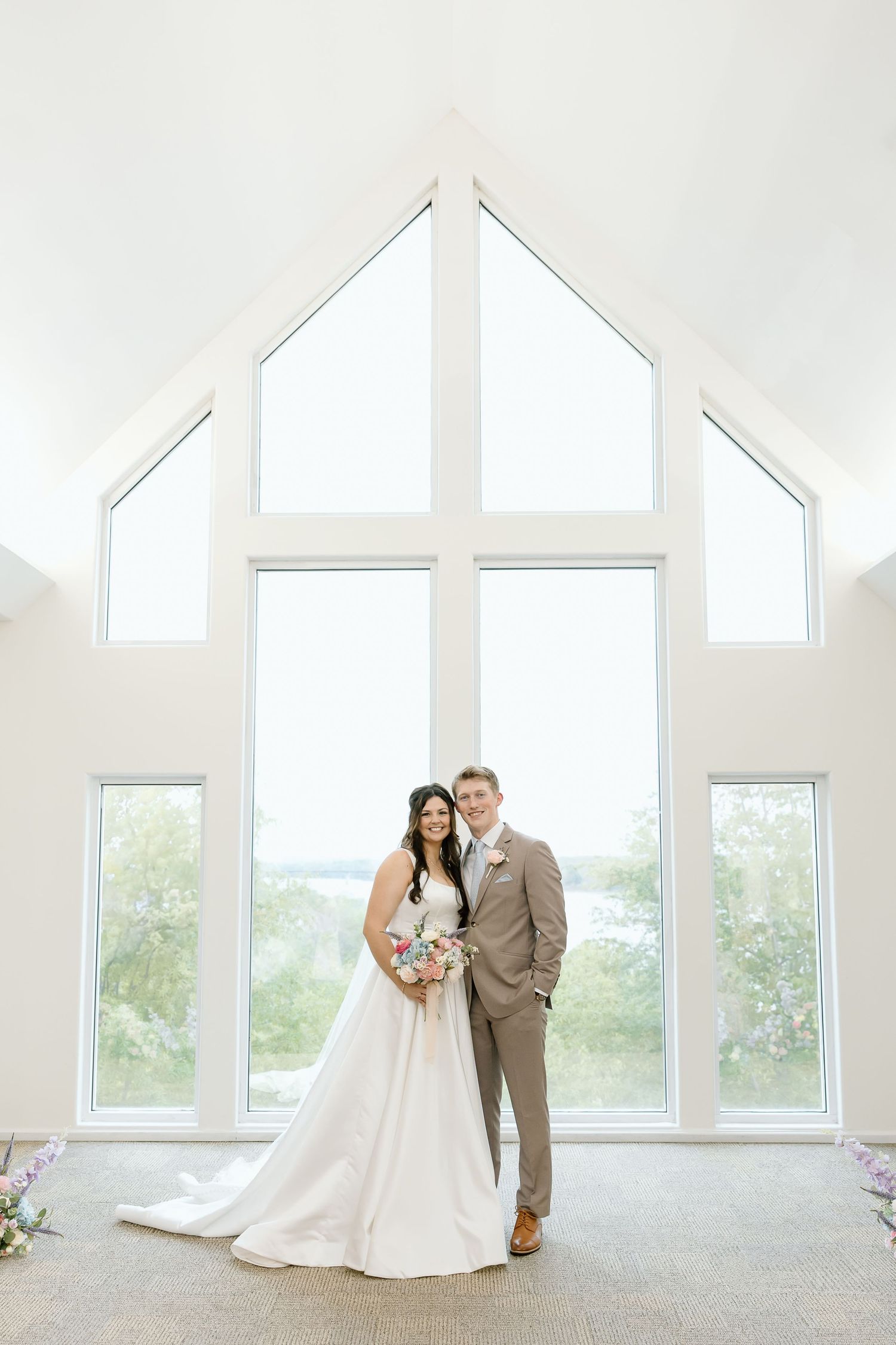 A couple in formal wedding attire poses in front of large cathedral windows in a bright white room.