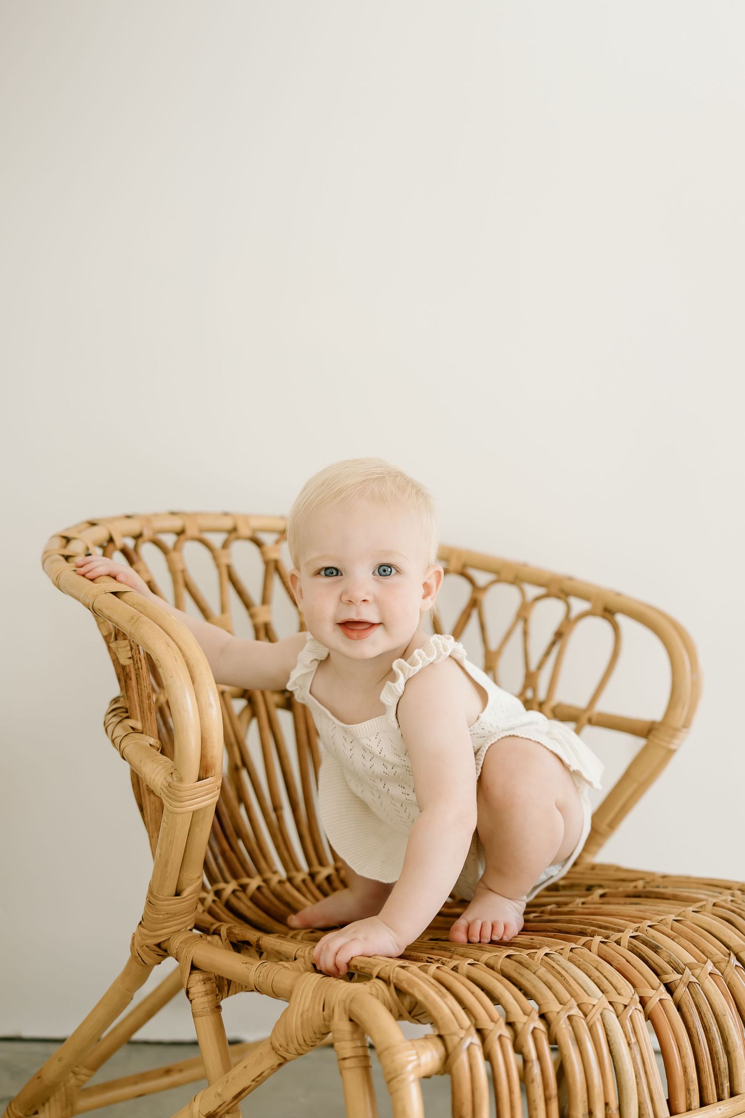 A baby sits on a vintage rattan chaise lounge chair against a neutral background.