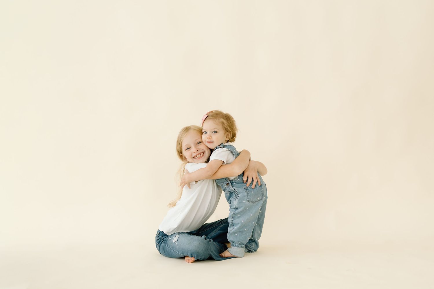 Two young children in light colored clothing sit close together against a white studio backdrop.
