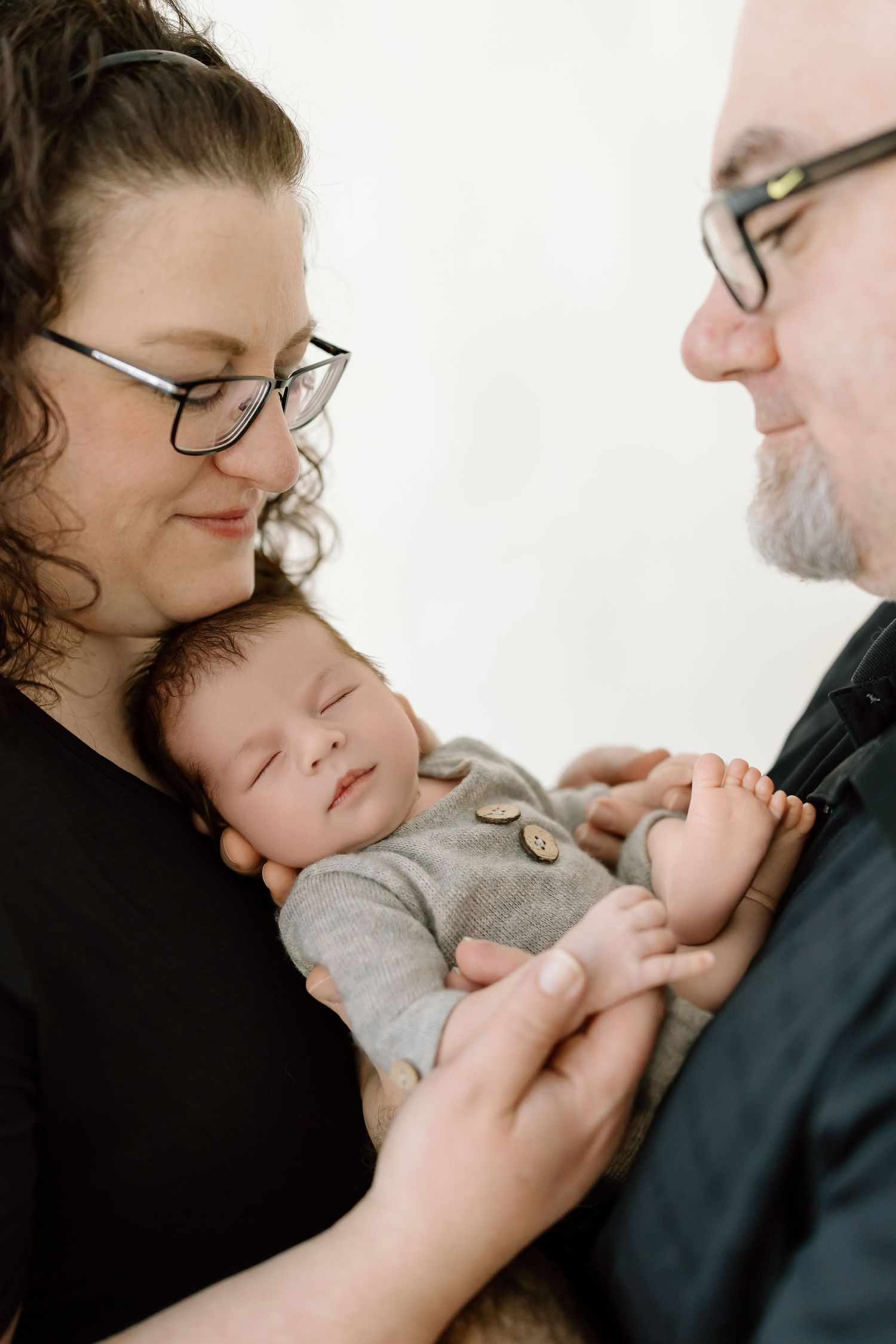 A sleeping newborn baby in gray onesie is cradled between parents wearing black clothing and glasses.