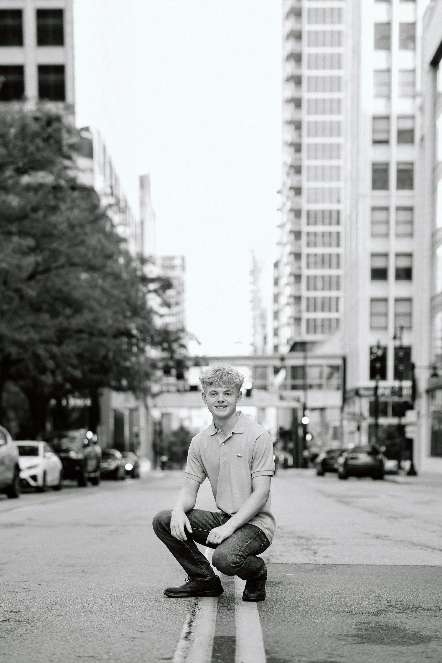 Black and white urban portrait of someone crouching in the middle of a city street with tall buildings in the background.