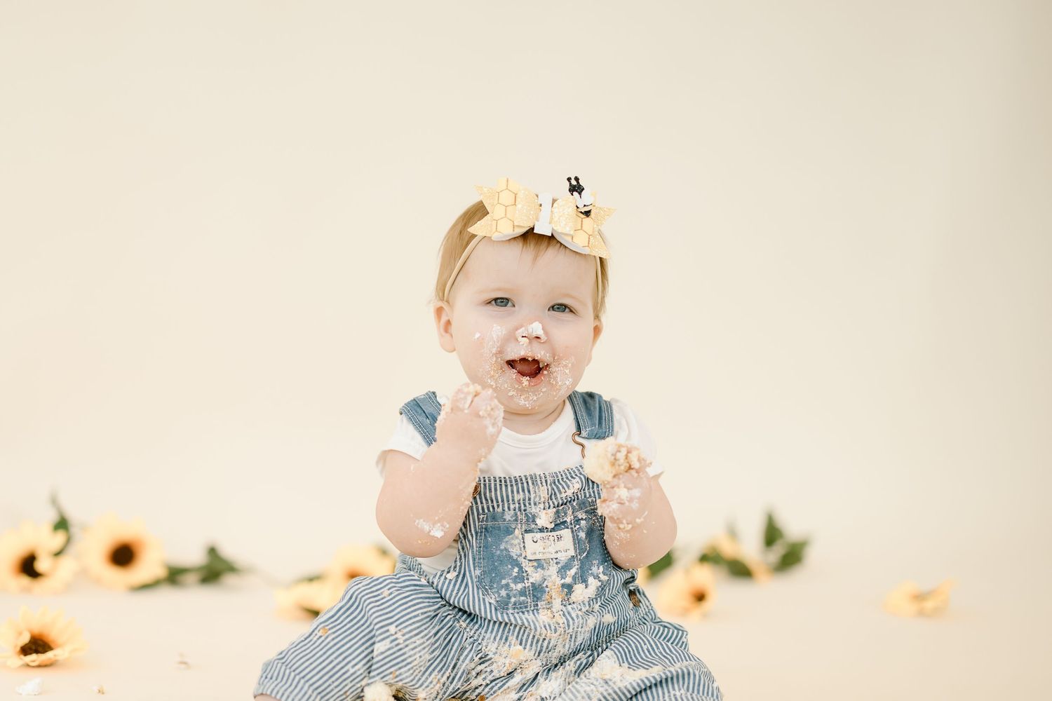 A young child wearing denim overalls sits and plays in a light-colored studio setting with sunflowers in the background.