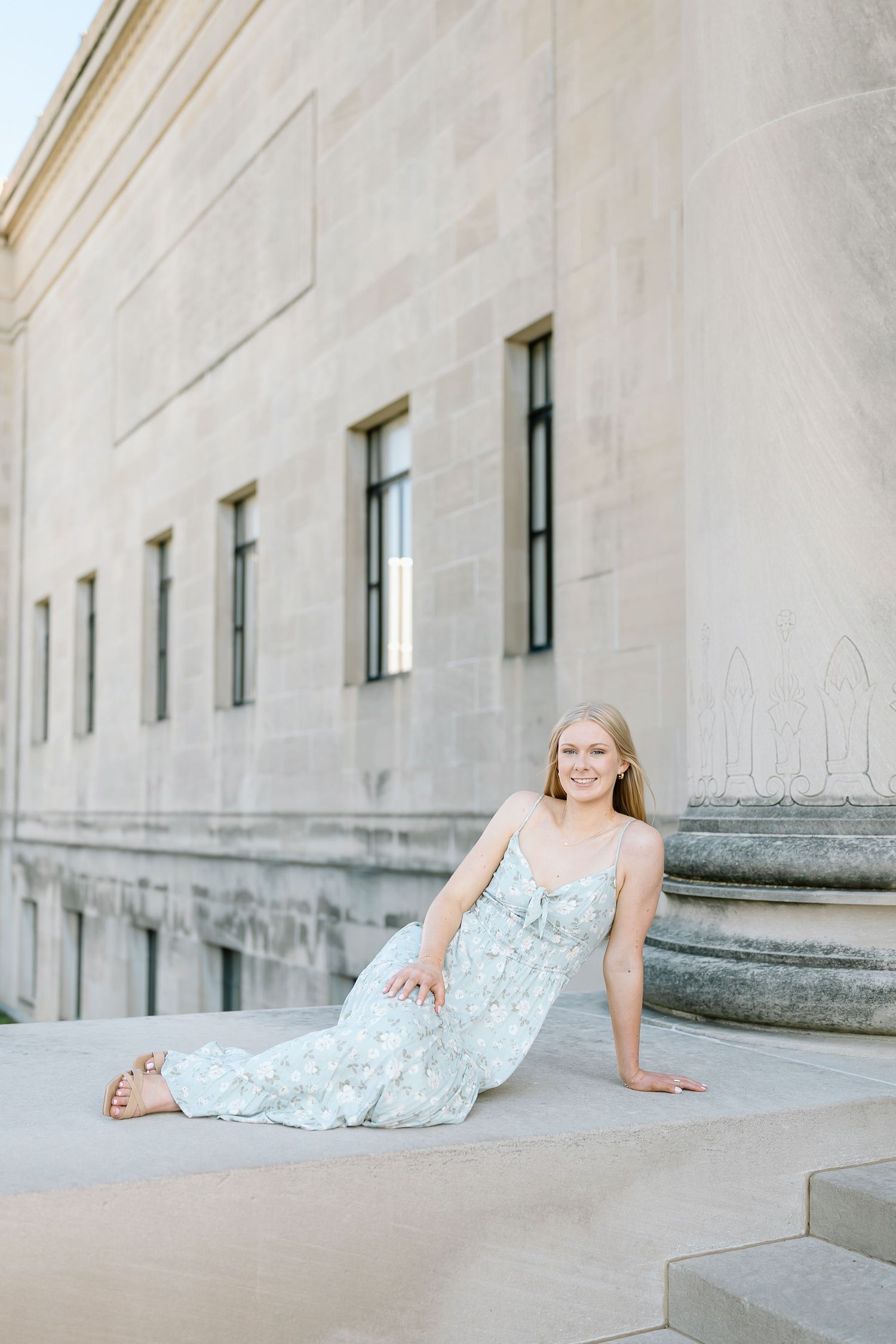 A woman in a floral dress reclines on concrete steps in an elegant outdoor architectural setting.