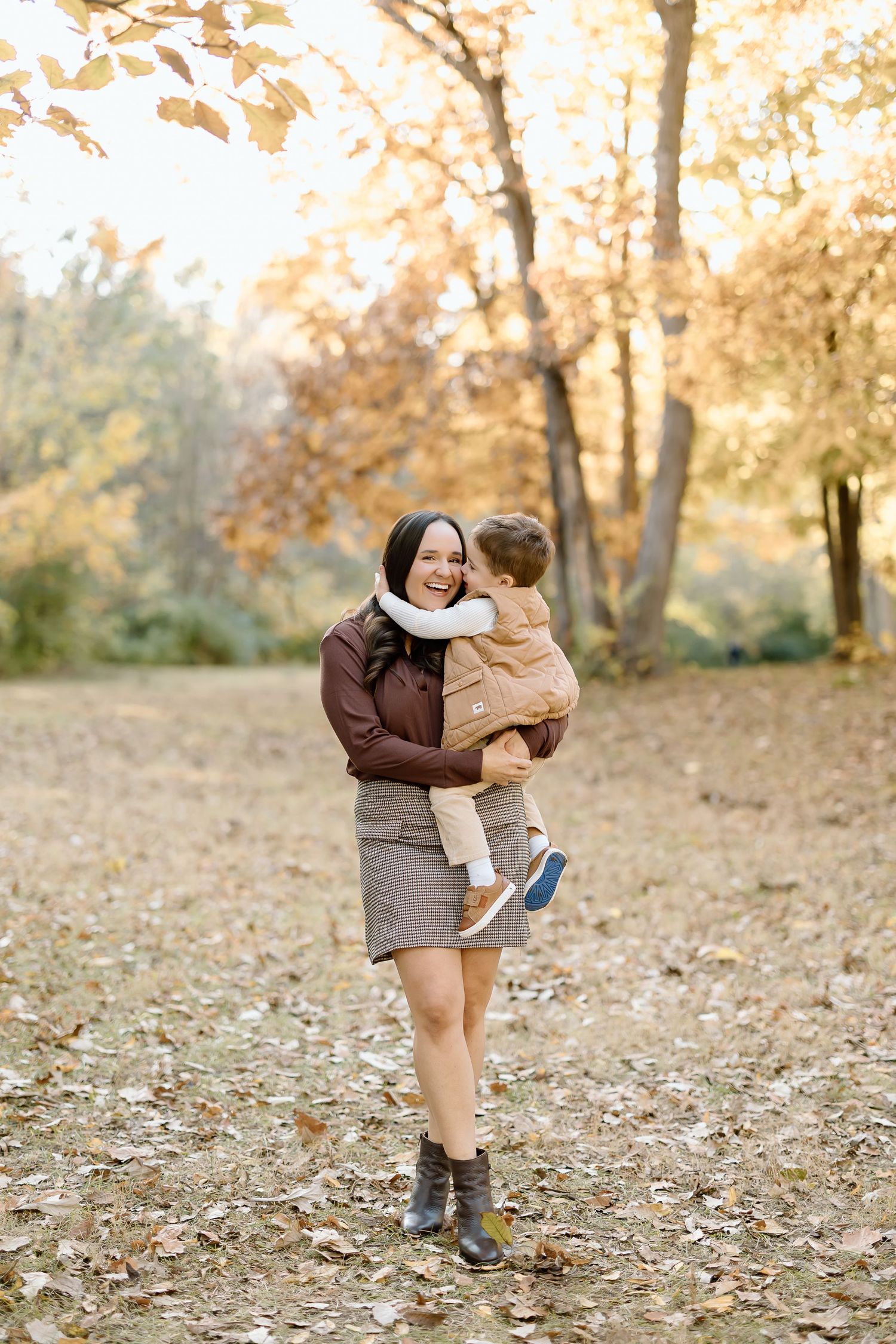 Two people share a tender moment on an autumn day surrounded by golden leaves and trees in a park setting.