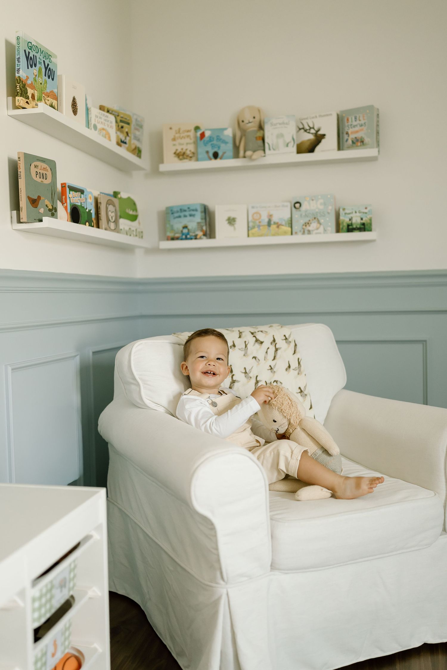 Cozy nursery corner with white armchair, floating bookshelves displaying children's books, and blue-gray wainscoting.
