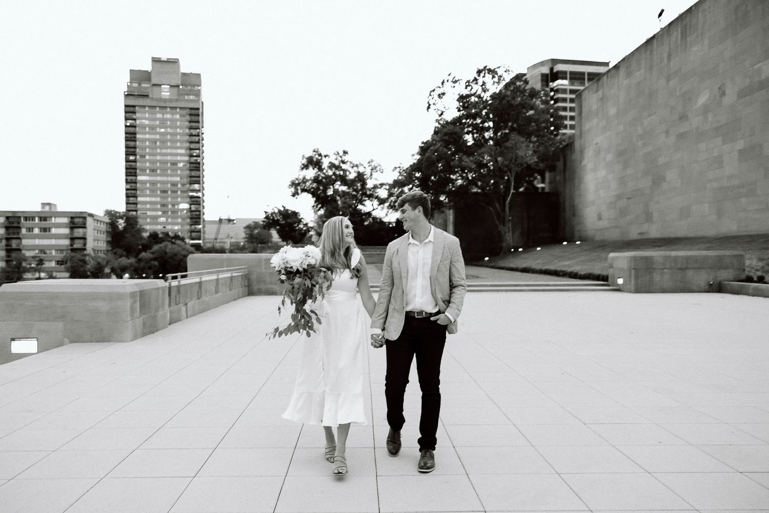 Two people walking together on a white concrete surface with tall buildings visible in the background.