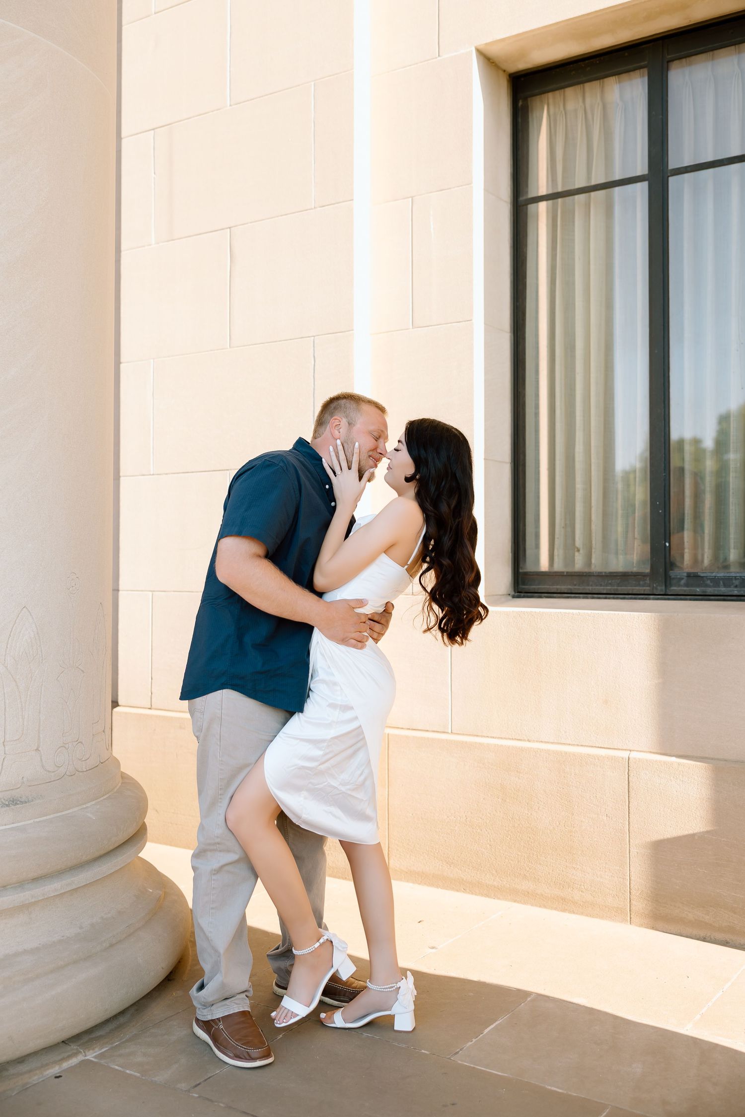 A romantic couple in white and navy outfits share a tender kiss against a beige stone wall in an outdoor setting.