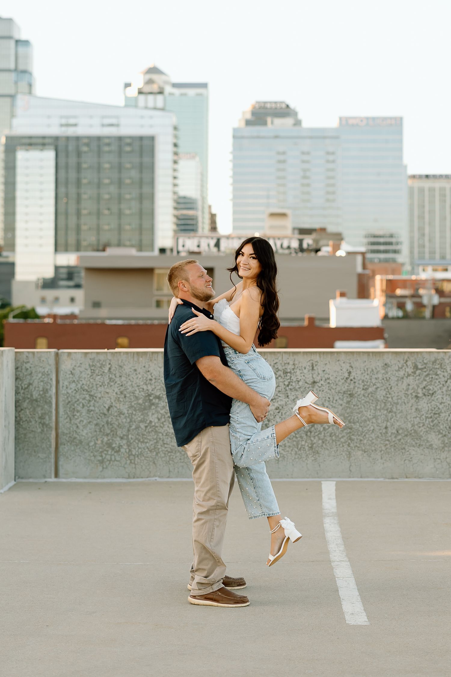 A couple shares a romantic dance and embrace on a rooftop with a city skyline in the background.