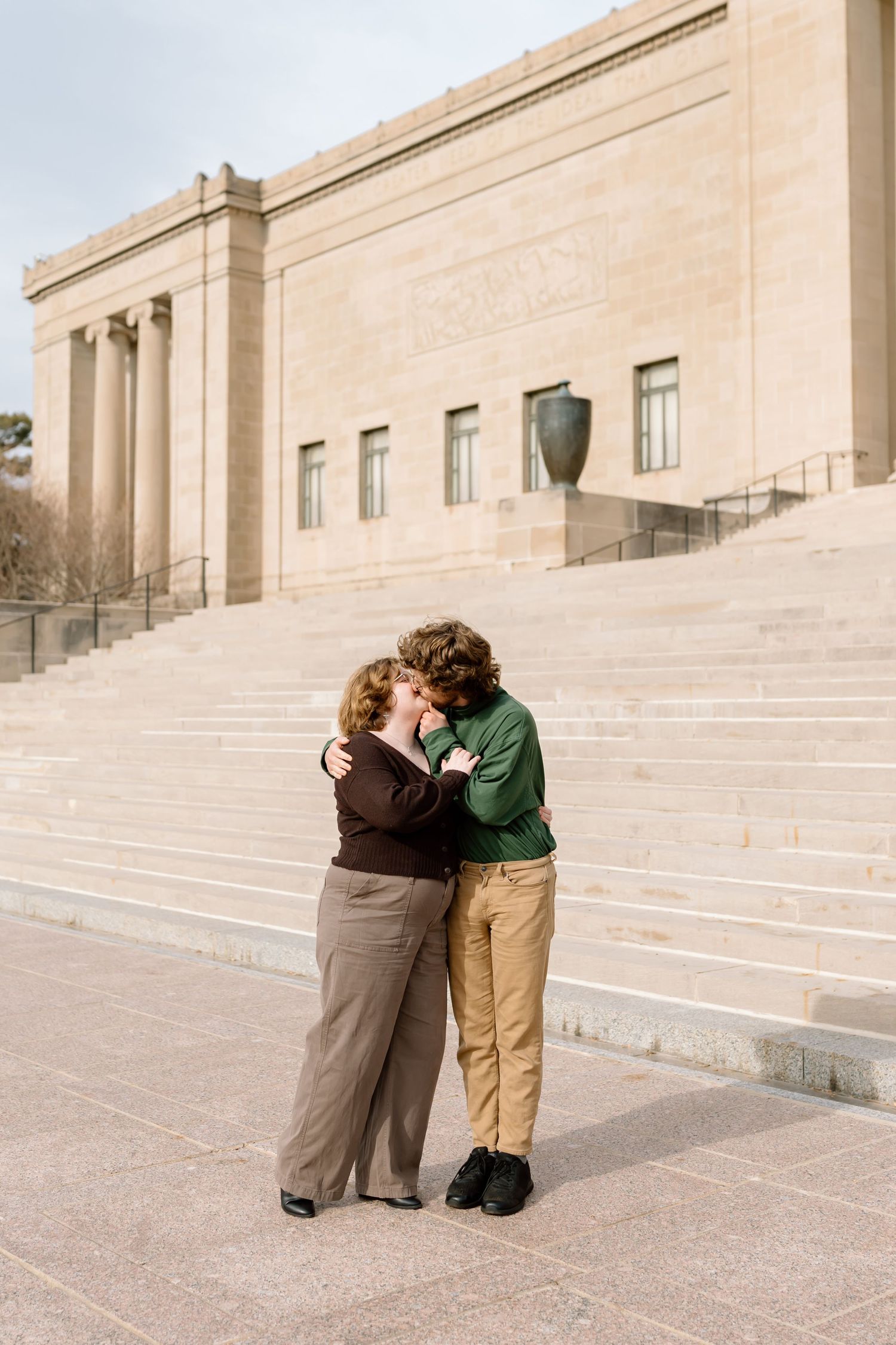 Two people embrace on wide stone steps of a grand building with beige limestone architecture.