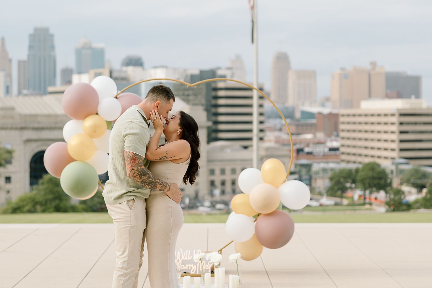 A couple holds pastel balloons on a rooftop with a cityscape backdrop during an engagement photoshoot.