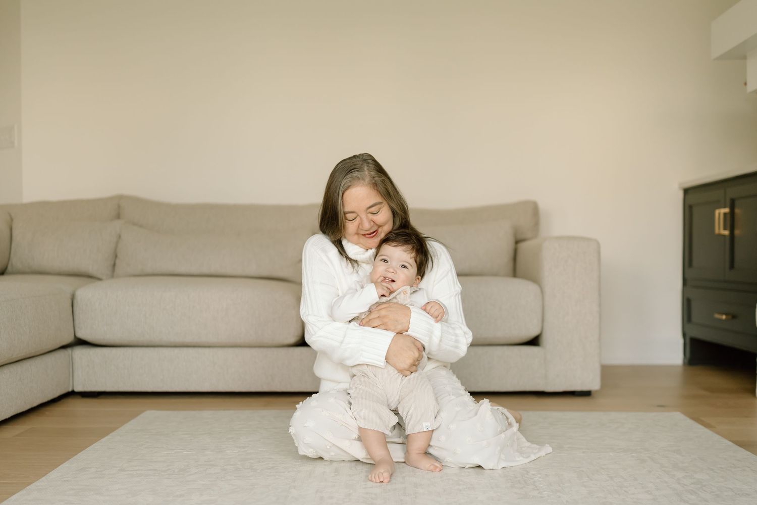 Two people in white clothing sit together on a beige carpet in a bright, minimalist living room setting.