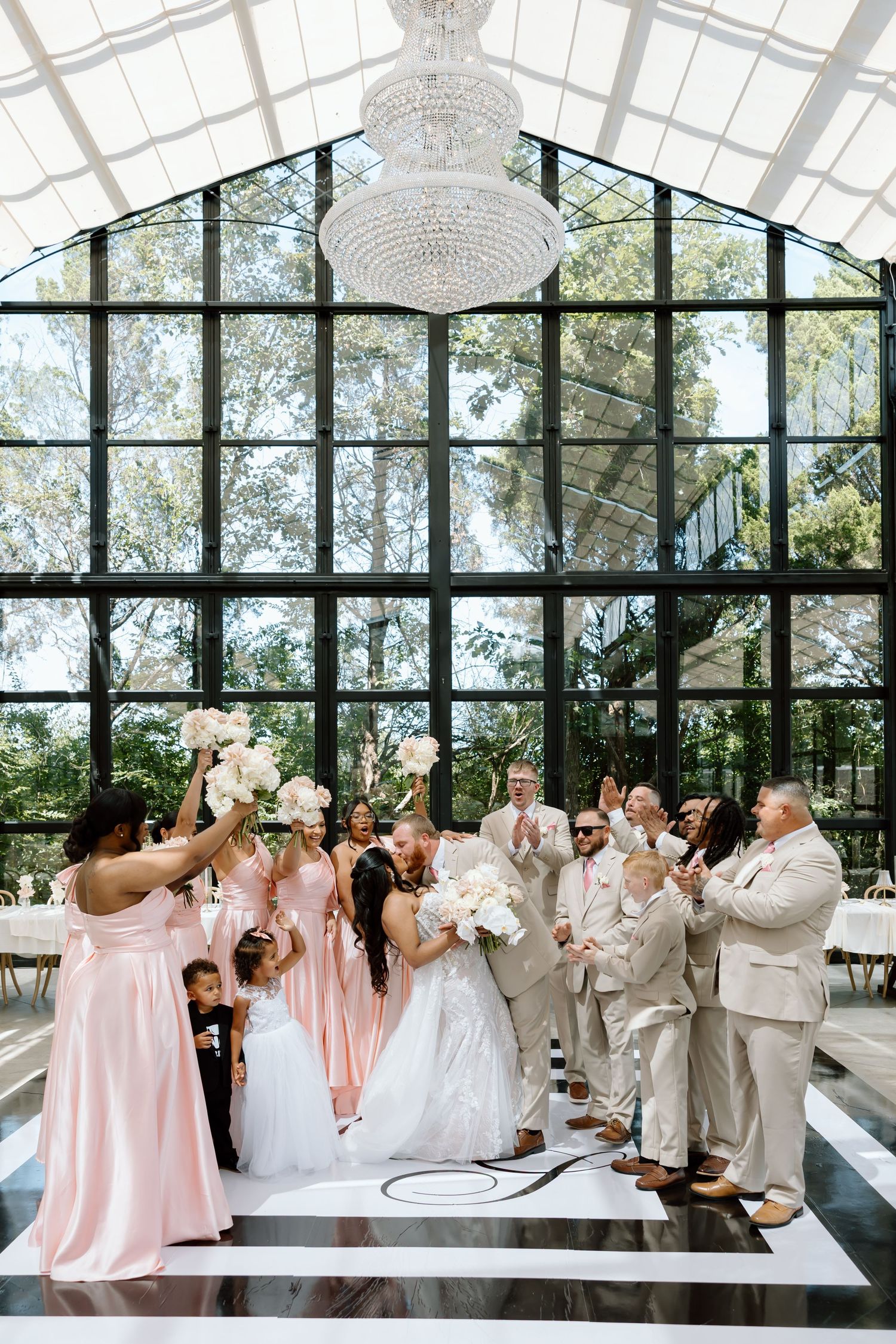 Two brides share a kiss surrounded by cheering wedding party in blush pink and tan suits inside a glass greenhouse venue.